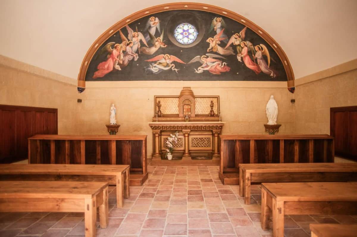 Interior of a small Catholic chapel with gilded altar, angel fresco, rose window, wood pews, and terracotta floor tiles