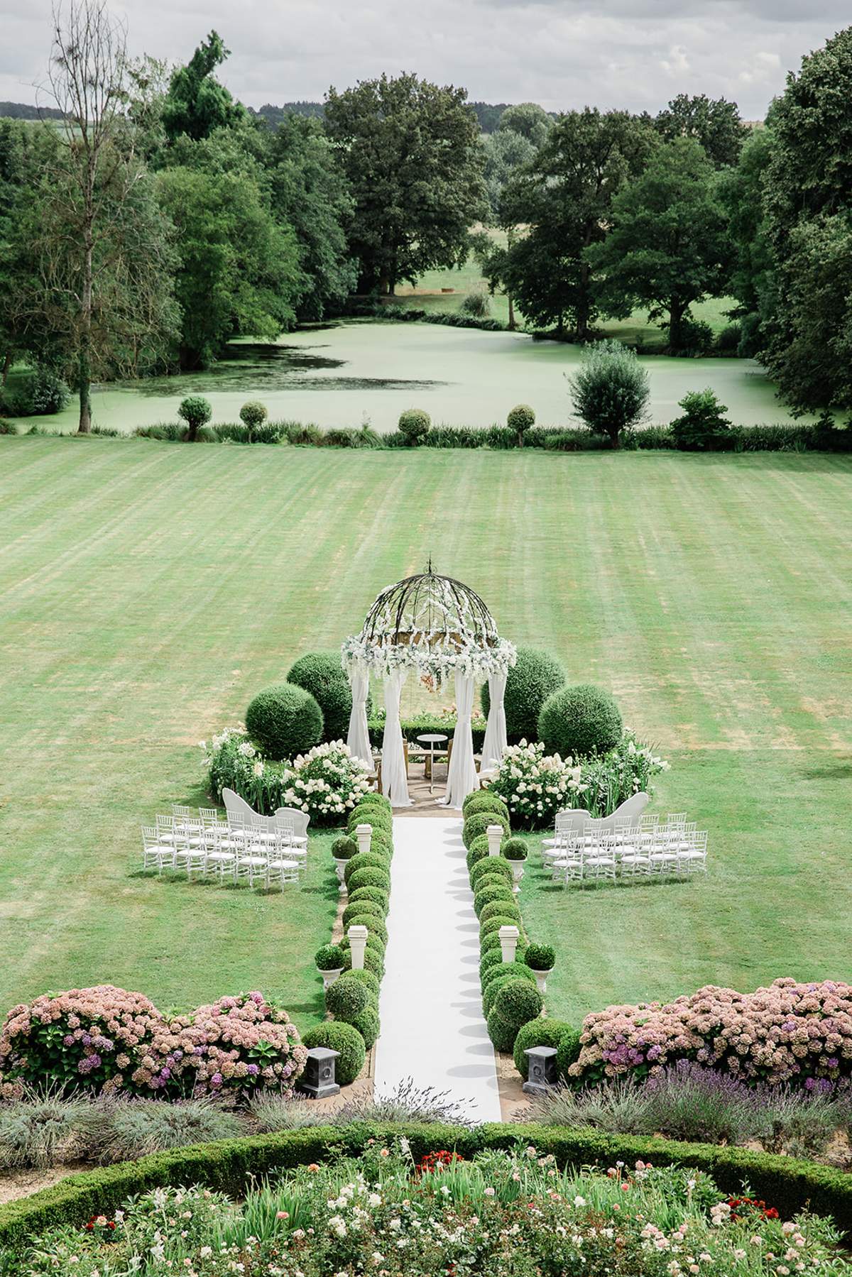 Aerial view of outdoor ceremony setup with white draped gazebo, floral arrangements, and chiavari chairs by ornamental lake
