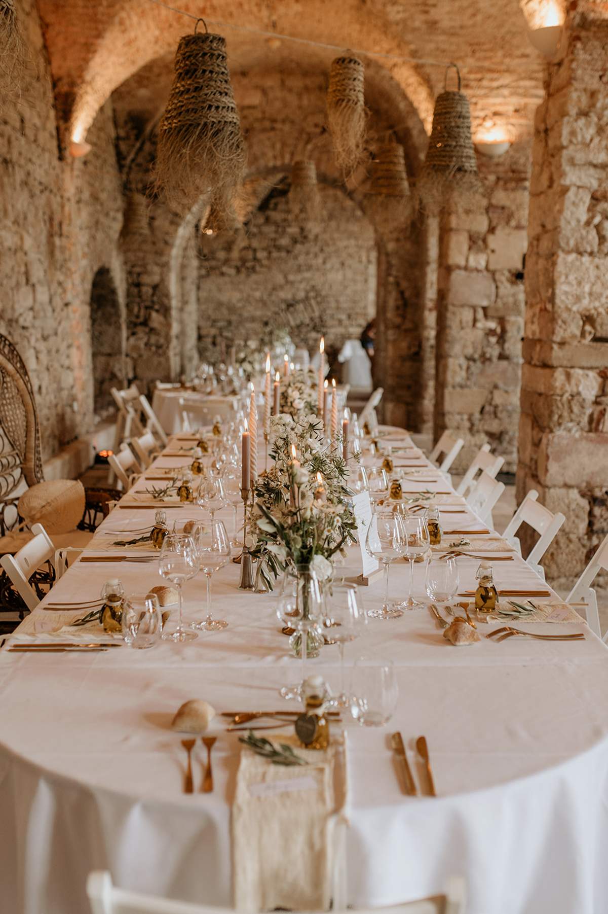 Vaulted stone reception with rattan pendants blush taper candles white floral bud vases olive oil favors and peacock chair