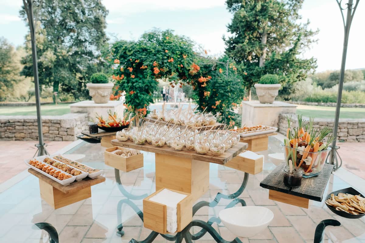 Canape station on wrought-iron table with tiered wood risers on stone terrace with formal garden beyond