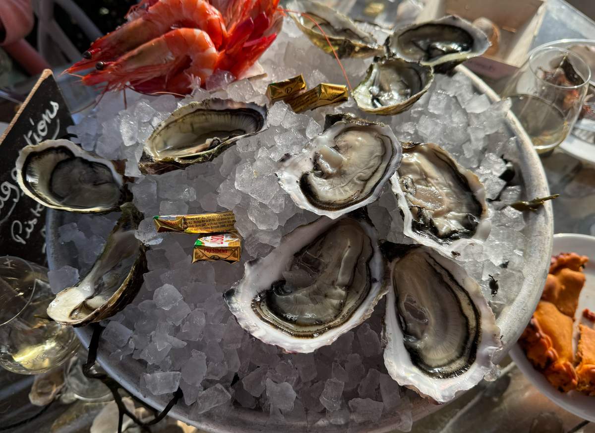 This image does not appear to be a wedding photograph. It shows a close-up overhead shot of a seafood platter at what looks like a casual outdoor dining setting, featuring freshly shucked oysters and whole prawns arranged on crushed ice on a round metal tray, with small individual Président butter packets scattered among the shells. Two glasses of white wine are partially visible at the edges, along with what appears to be a chalkboard menu sign and a partial view of a crab dish. This image is not suitable for a wedding photography content library.