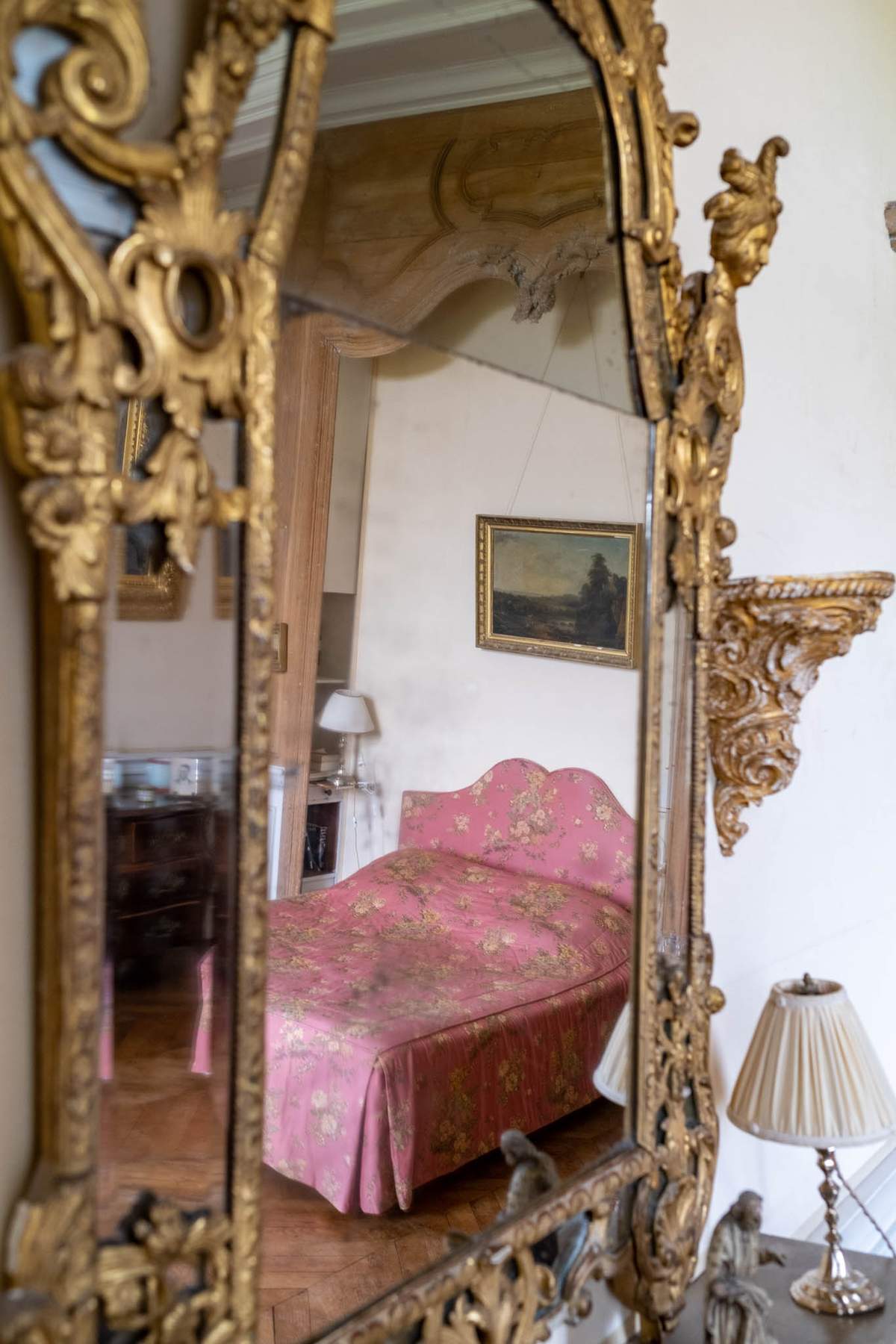 Ornate gilt Baroque mirror reflecting pink floral daybed landscape painting and ceiling moldings in chateau interior