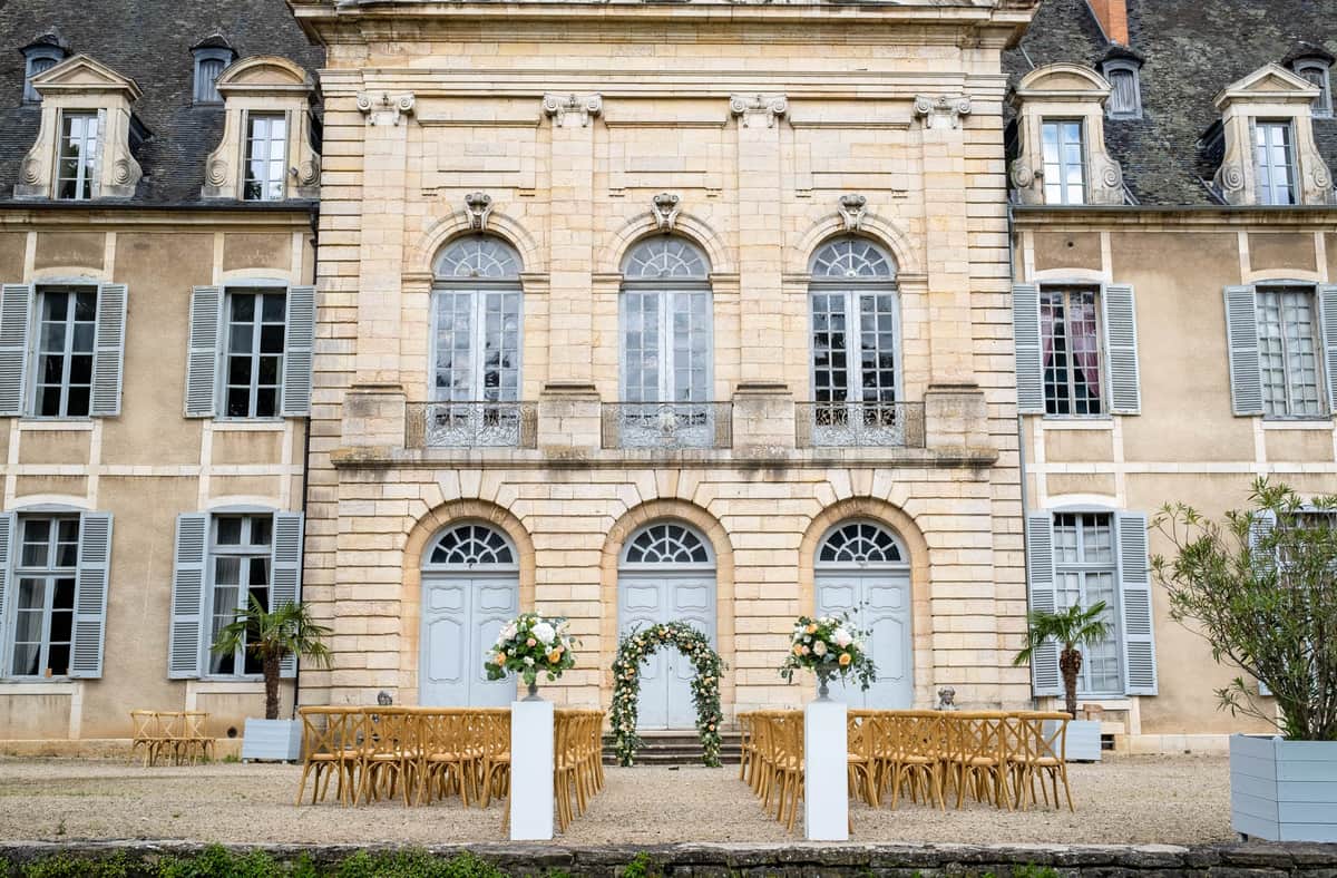 Outdoor ceremony setup with cross-back chairs and circular floral arch before chateau facade