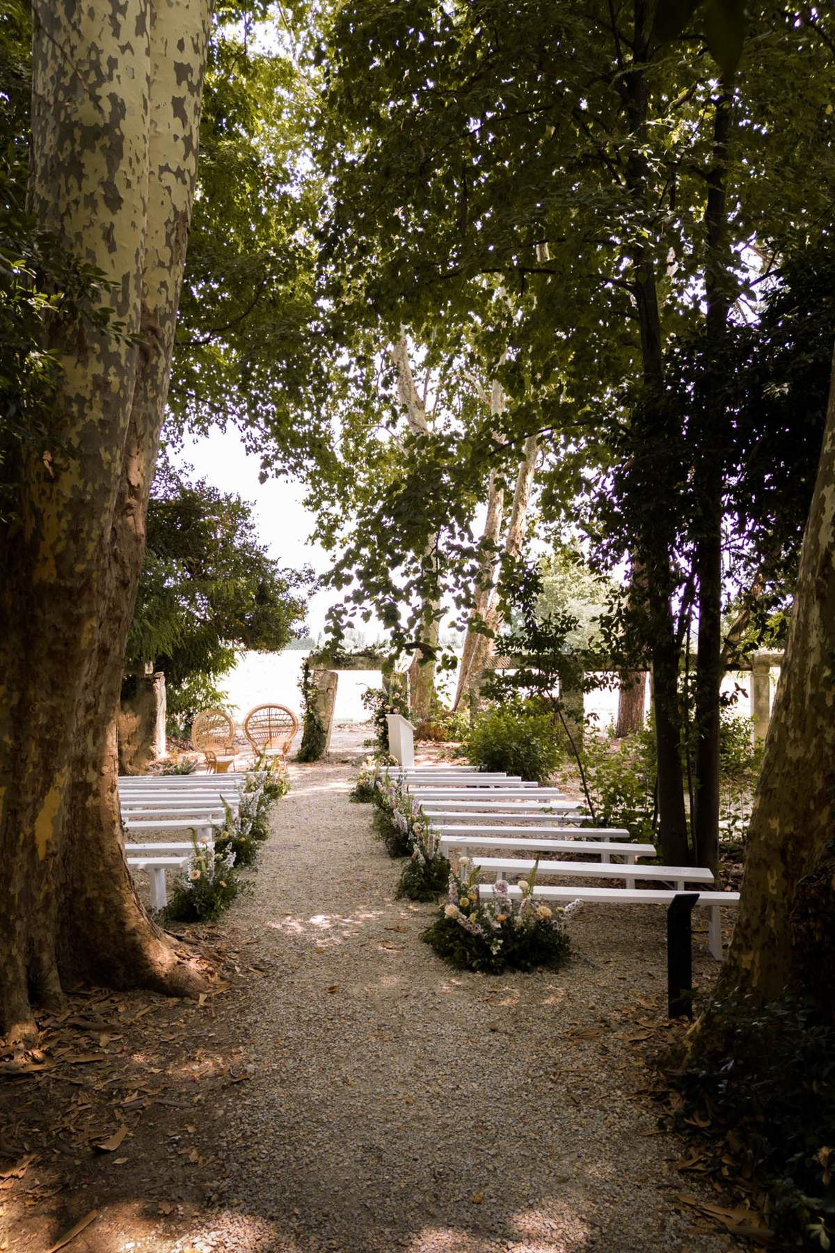 An outdoor ceremony setup photographed before guests arrive, captured as a wide shot looking down the aisle toward the altar. The ceremony space is set within a tree-lined gravel path, with white wooden benches arranged in two rows on either side of the aisle. Low floral arrangements in soft white, cream, and pale yellow tones are placed at the end of each bench row along the aisle. At the far end, two large round rattan peacock chairs are positioned near the altar area, which features a greenery-draped stone structure or pergola. The overall styling is boho with natural, organic elements, using white painted furniture against the gravel and tree canopy. A body of water is visible in the background through the trees. Potential venue feature image.