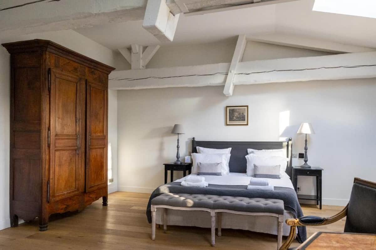 Interior shot of a guest bedroom, likely within a French château or manor house venue. The room features exposed white-painted timber ceiling beams, light oak hardwood flooring, and white walls. The bed has a dark charcoal iron frame dressed in white and grey linen with folded towels placed on top, flanked by two dark bedside tables with ivory lamp shades. A large antique French armoire in warm walnut wood stands to the left, and a grey tufted upholstered bench sits at the foot of the bed. A leather chair is partially visible in the foreground right. The styling mixes antique French provincial furniture with a clean, neutral grey and white palette. Wide interior shot. Potential venue feature image.