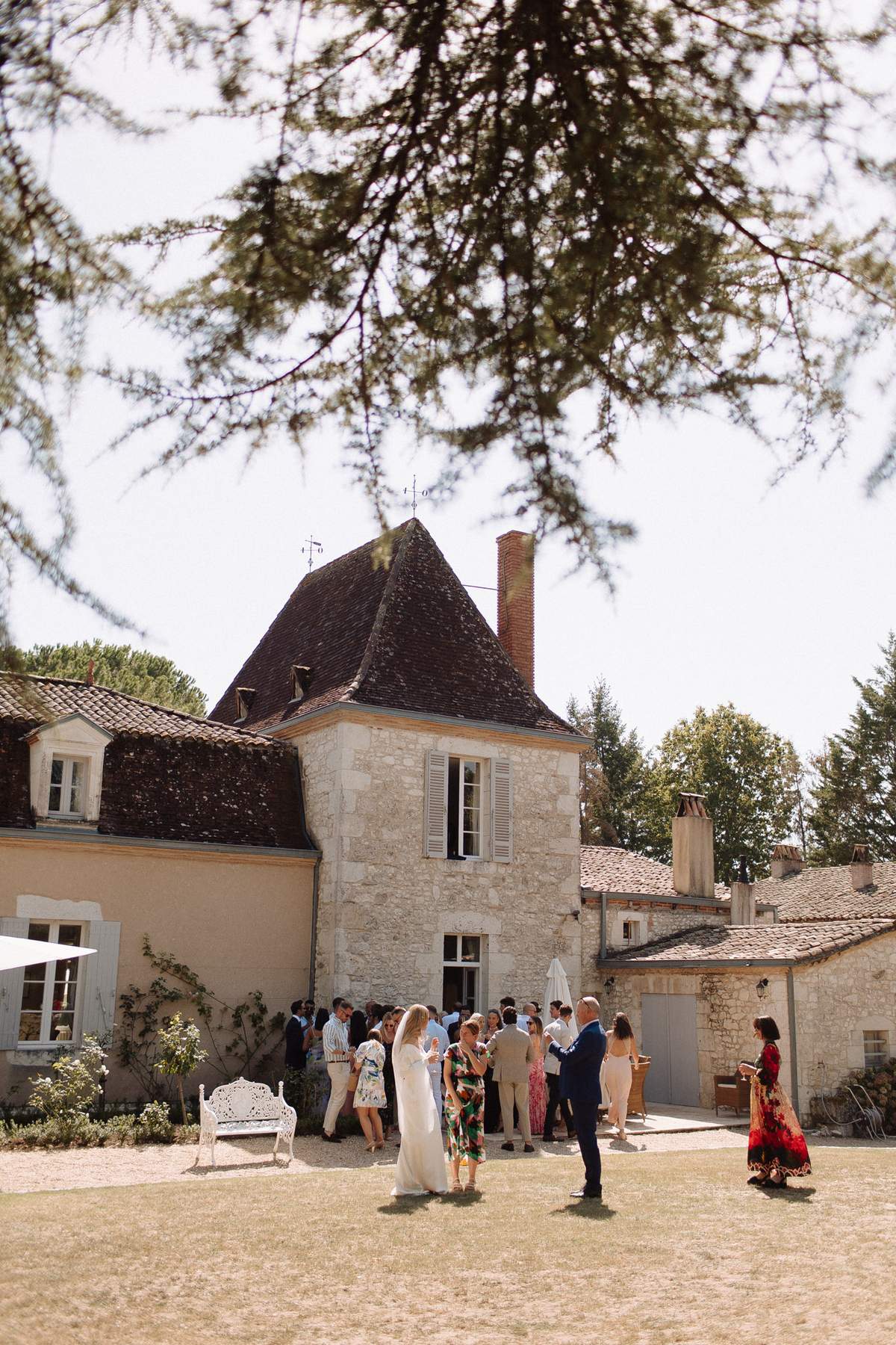 An outdoor cocktail hour taking place in the courtyard of a French stone manor house featuring a distinctive pointed tower with a dark slate roof, white shuttered windows, and terracotta tile roofing on the adjoining wings. Approximately 20–25 guests are mingling in small groups across the courtyard, with the bride visible in a white floor-length dress near the center foreground, accompanied by a groom in a navy suit. Guests are dressed in a mix of colorful summer attire including a bold green floral dress and a red patterned maxi dress. A white ornate garden bench is positioned to the left side of the courtyard. The shot is a wide, slightly elevated perspective framed by overhanging tree branches in the upper portion of the frame, captured in bright midday light. Potential venue feature image.
