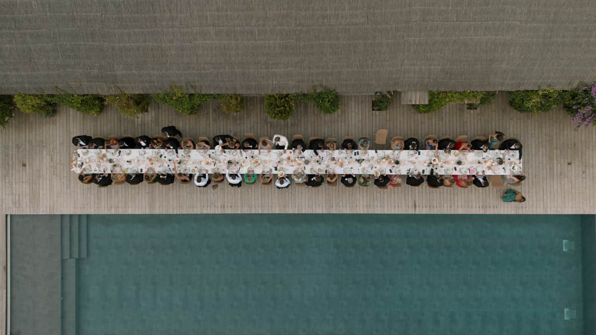 Aerial view of wedding reception dinner along a long table on wooden deck beside a swimming pool