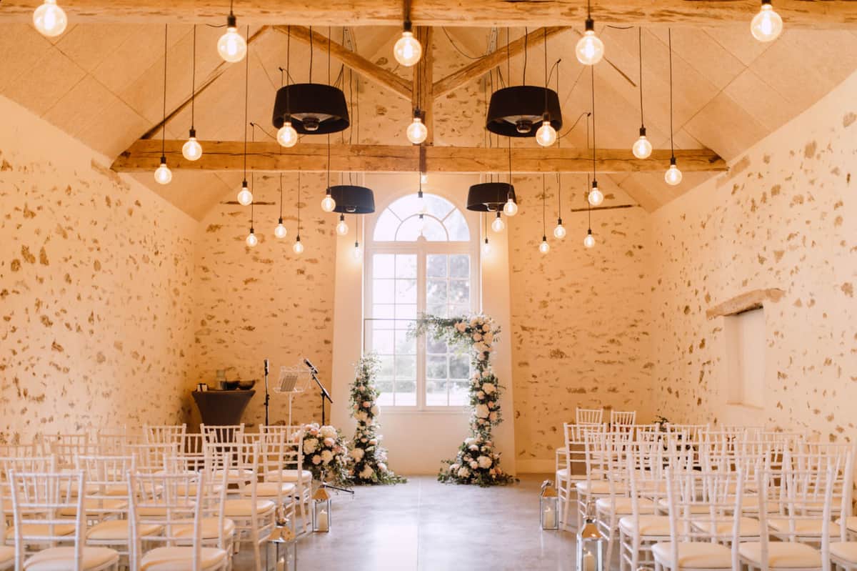 Indoor ceremony space with circular blush and ivory floral arch white Chiavari chairs and Edison pendant lights