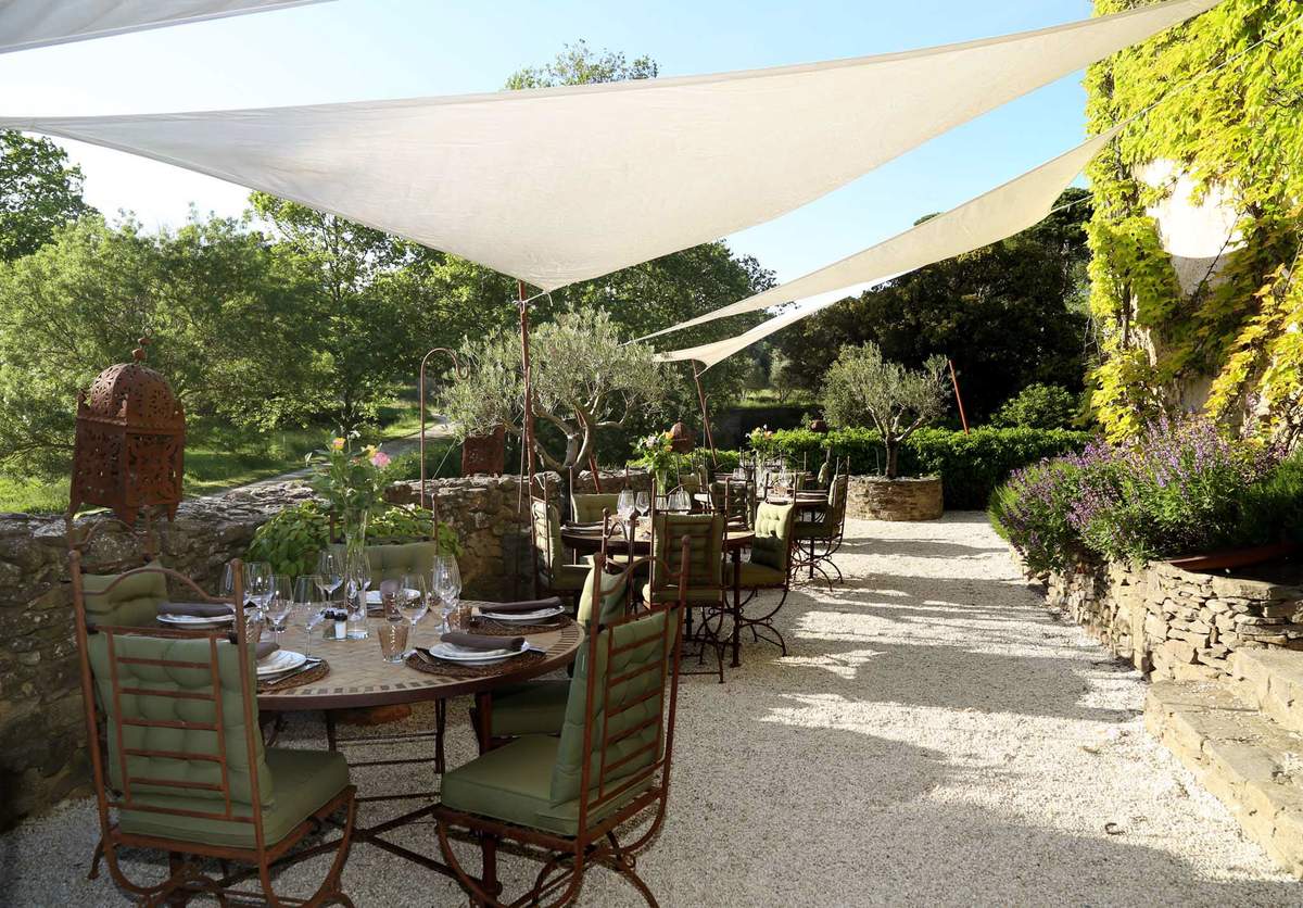 An outdoor reception dining setup on a gravel terrace, shaded by large white triangular sail canopies stretched overhead. Multiple round iron tables are set for dining with wine glasses, white folded napkins on woven charger plates, and small floral centerpieces with pink blooms. The wrought-iron chairs have sage green cushioned seats and backs. Decorative rust-patinated Moroccan-style lanterns are placed on the tables and mounted on curved iron stands throughout the space. The terrace is bordered by low dry-stone walls with lavender plantings and trimmed hedges along the right side. No people are present in the image. The overall styling is Provençal with a relaxed, rustic-Mediterranean aesthetic. Wide shot taken from a low angle looking down the length of the terrace. Potential venue feature image.