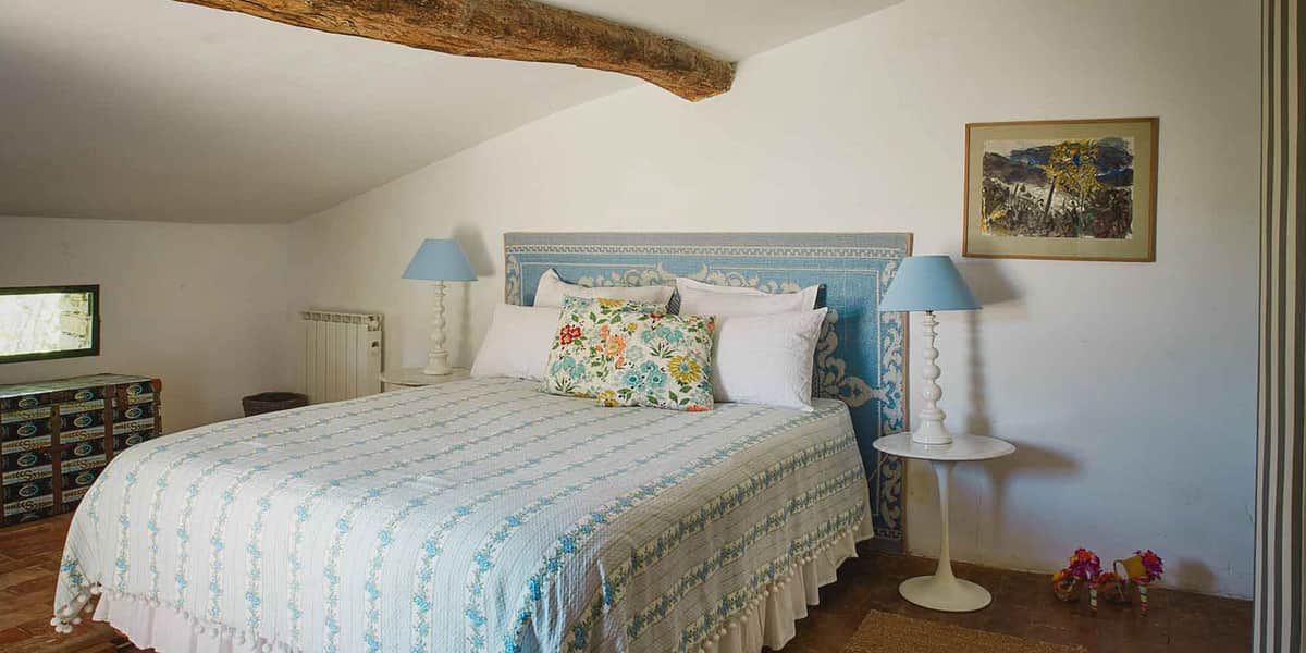 An interior shot of a guest bedroom at what appears to be a French country property, likely used as accommodation for a wedding event. The room features a double bed with a blue-and-white tiled or embroidered headboard, dressed in a white and light blue floral quilted bedspread with pom-pom trim and multiple white pillows, including a multicolored floral accent cushion in blue, yellow, green, and red. Two matching white candlestick-style lamps with pale blue shades sit on either side of the bed on white side tables. The room has a vaulted ceiling with an exposed rustic wooden beam, terracotta tile flooring, and white walls with a framed artwork print above the right nightstand. A vintage trunk is visible in the background left corner. The overall styling is rustic-provincial with blue and white tones. Wide interior shot. Potential venue feature image.