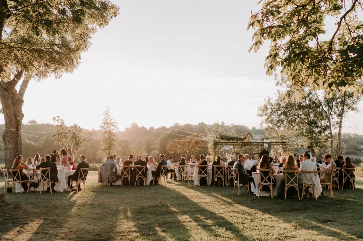An outdoor wedding reception dinner is underway on a large lawn, with approximately 60–80 guests seated at multiple round tables covered in white linens. The tables are surrounded by natural wood cross-back chairs, consistent with a rustic-country styling theme. String lights are strung overhead between the trees, and a greenery-covered pergola structure is visible in the background center, draped with climbing foliage. The scene is lit by warm late-afternoon golden hour light, casting long shadows across the lawn and bathing the entire setting in an amber glow. Guests are dressed in a mix of formal and semi-formal attire, including dark suits and dresses in black, red, and neutral tones. The wide-angle shot is taken from ground level looking across the lawn toward the open countryside beyond.