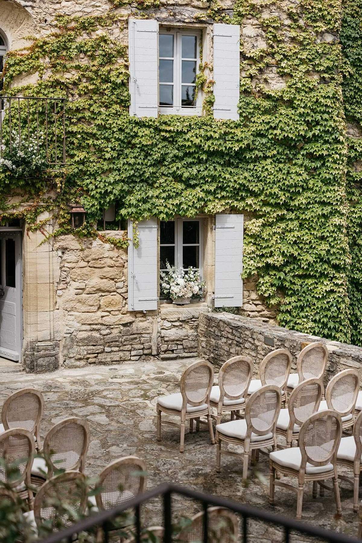 An outdoor ceremony setup in a stone courtyard in front of a French château or manor house facade with pale grey shuttered windows. Rows of French Louis-style chairs with cane backs and cream upholstered seat cushions are arranged in a curved seating configuration facing the building. A floral arrangement of white and cream blooms is displayed on the lower windowsill as a ceremony backdrop decoration. The overall decor palette is neutral — natural wood tones, cream, and soft grey — consistent with a classic French country styling theme. The image is a wide shot capturing both the seating arrangement and the architectural facade. Potential venue feature image.
