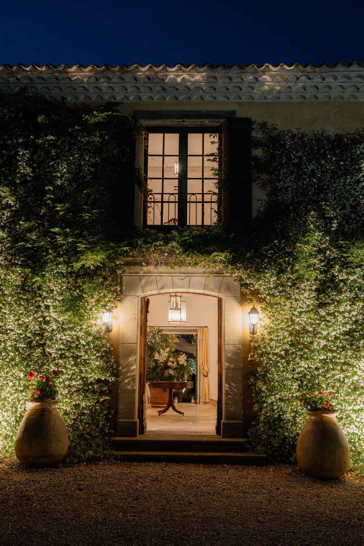 A nighttime exterior shot of a historic mas or domaine-style venue, showing the main entrance facade lit by warm wall-mounted lanterns and a hanging pendant light framing the open arched doorway. The two-story stone building is heavily covered in climbing vines with small white blooms, illuminated by uplighting that highlights the texture of the greenery against the pale render. Two large terracotta urns planted with red geraniums flank the entrance on a gravel forecourt. Through the open double doors, a warmly lit interior is visible with a dark wood console table, cream curtains, and white floral arrangements. A tall iron-framed window on the upper floor is backlit from within, revealing a pendant light inside. The wide architectural shot is taken straight-on, centered on the doorway. Potential venue feature image.