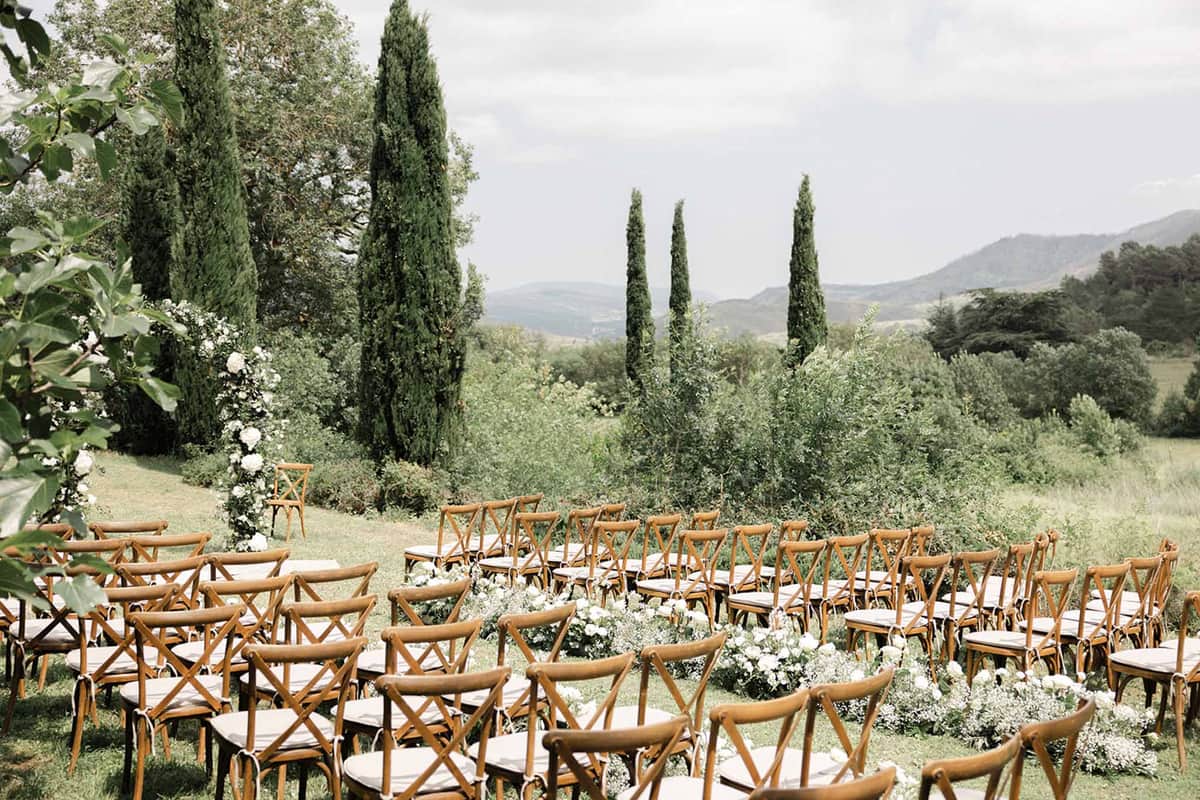 Outdoor ceremony setup with crossback chairs and cream cushions on lawn, white floral aisle markers and cypress tree backdrop