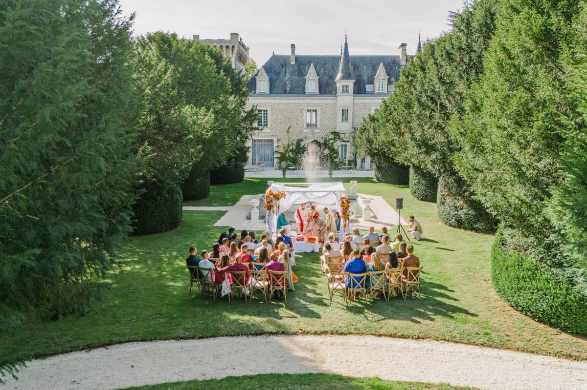 Elevated view of Hindu wedding ceremony under white mandap with orange florals in French chateau garden with seated guests