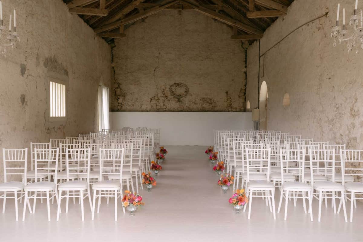 Rustic barn ceremony setup with white chiavari chairs and orange and pink floral aisle arrangements