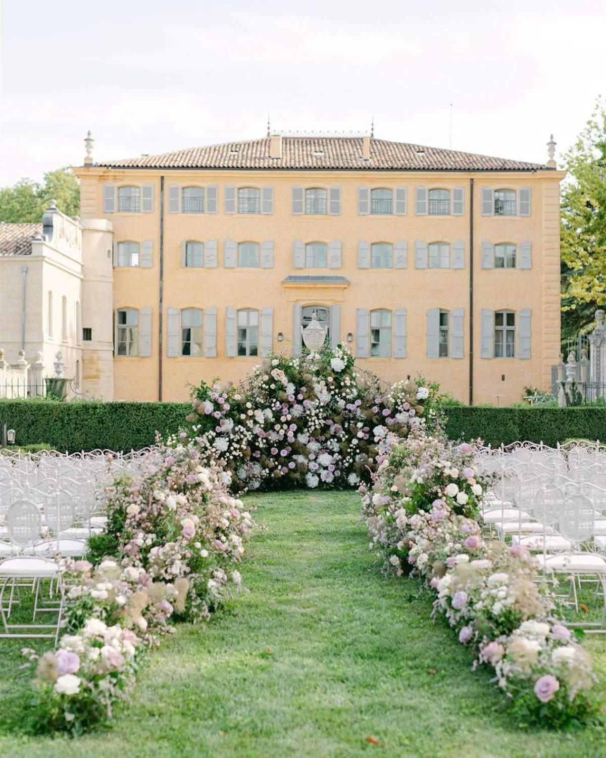 An outdoor wedding ceremony setup on a formal lawn in front of a three-story French château with warm ochre-yellow render and pale blue-grey shutters. The aisle is flanked by two curved floral arches and low ground-level floral runners, all composed of blush pink roses, mauve and lilac roses, ivory white blooms, pampas grass, and trailing greenery. At the altar, a large dense floral installation surrounds a white stone urn pedestal, continuing the same blush, mauve, and ivory palette with abundant foliage. White ghost-style acrylic chairs with white cushions are arranged in rows on either side of the aisle, accommodating what appears to be 60–80 guests. The overall styling is romantic and garden-inspired with a soft pastel color palette. Wide establishing shot taken from the entrance of the aisle looking toward the altar and château facade. Potential venue feature image.