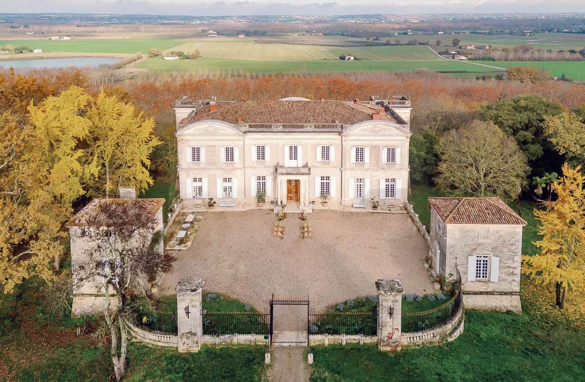 Aerial drone shot of a French classical chateau with cream limestone facade, terracotta tile roof, and white shuttered windows across two stories. The property features a symmetrical gravel forecourt flanked by two smaller stone outbuildings, with a wrought-iron entrance gate and low stone boundary wall at the front. A small floral arrangement or decorative display is visible in the center of the courtyard, suggesting event setup. The chateau is surrounded by mature trees with autumn foliage in gold and amber tones, with open agricultural fields and a small body of water visible in the background. Potential venue feature image.
