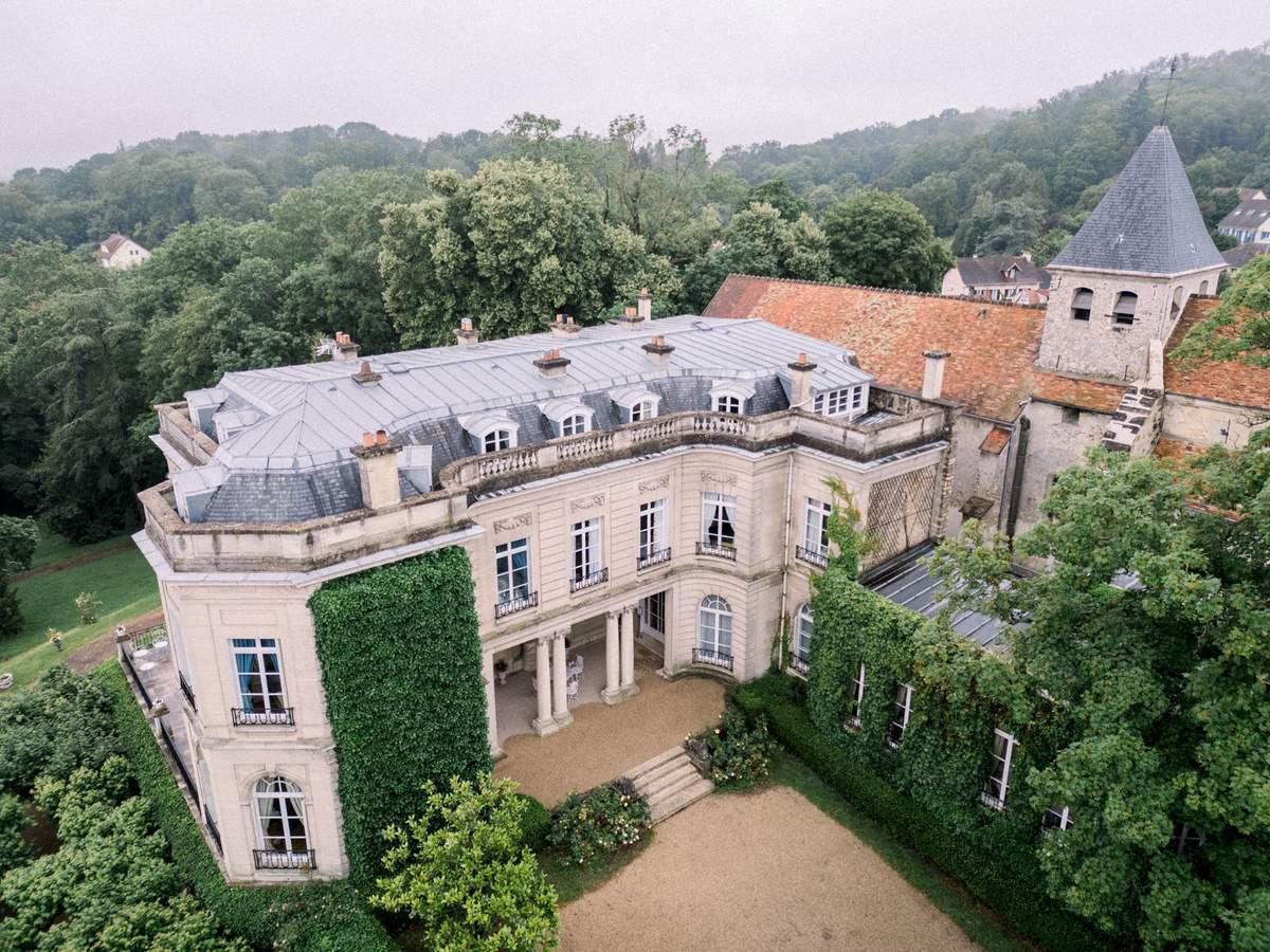 Aerial drone view of classical French chateau with mansard roof, ivy-covered facade, and adjacent chapel tower