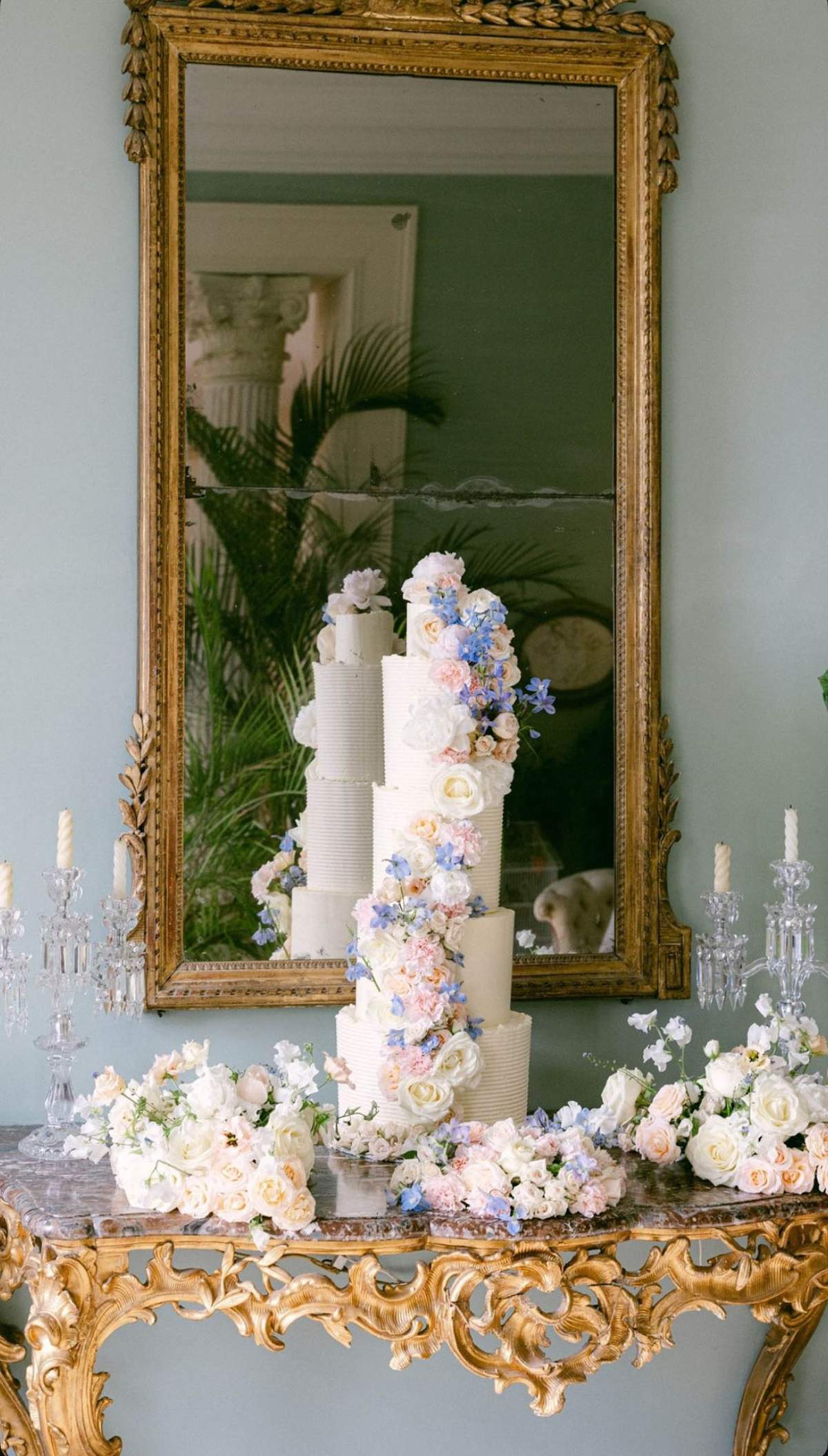 Five-tier ribbed buttercream wedding cake with cascading pink roses and blue delphinium on a gilt console table