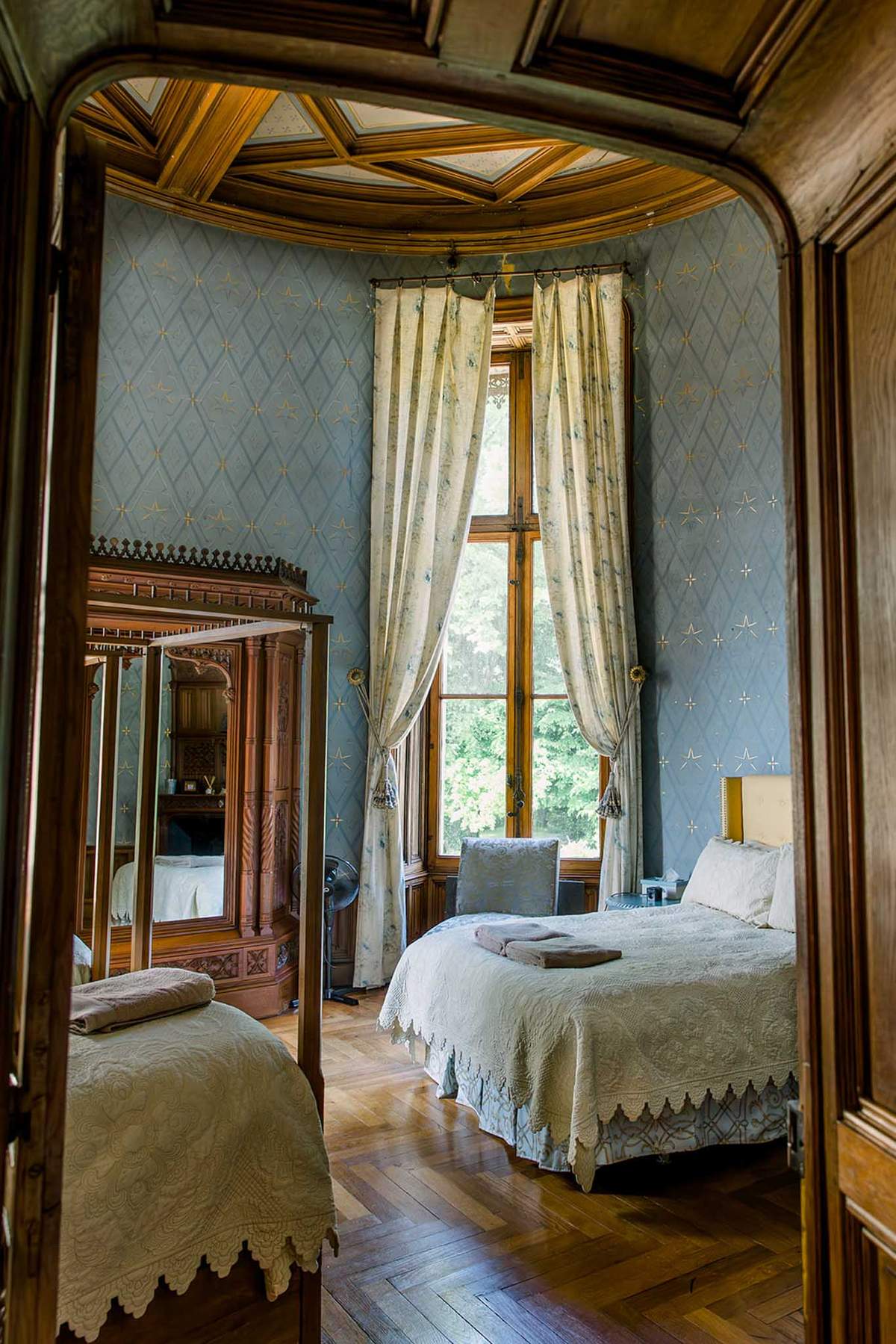 Chateau bedroom with powder blue wallpaper, coffered ceiling, mirrored armoire, and lace-trimmed beds