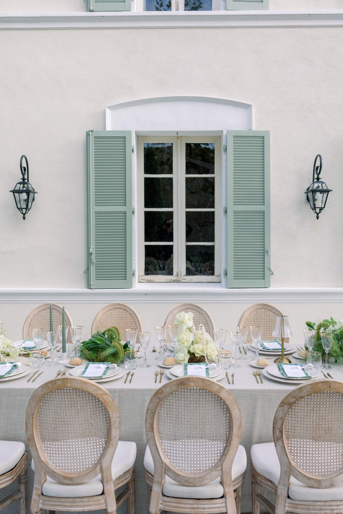 Long table with sage candles, ivory roses, and cane-back chairs against white facade with green shutters