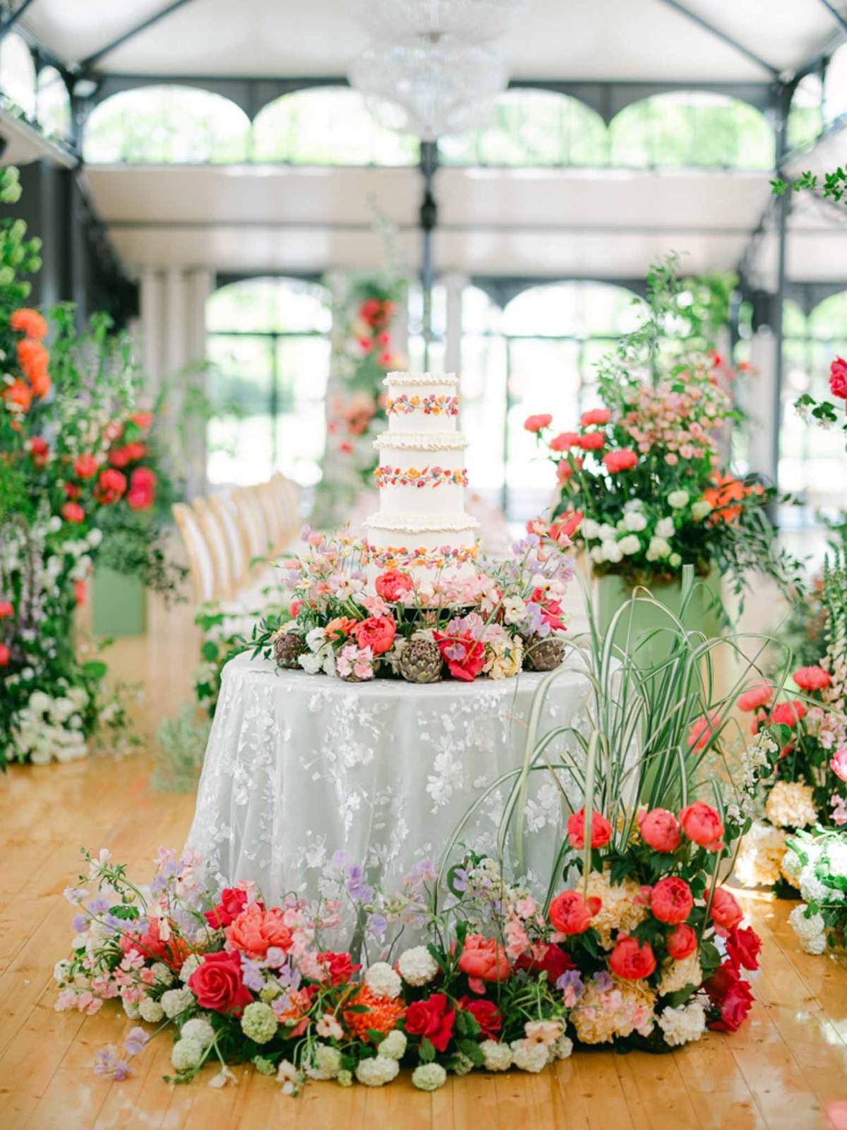 Four-tier white wedding cake with painted florals surrounded by coral peonies and red roses in a glass orangery