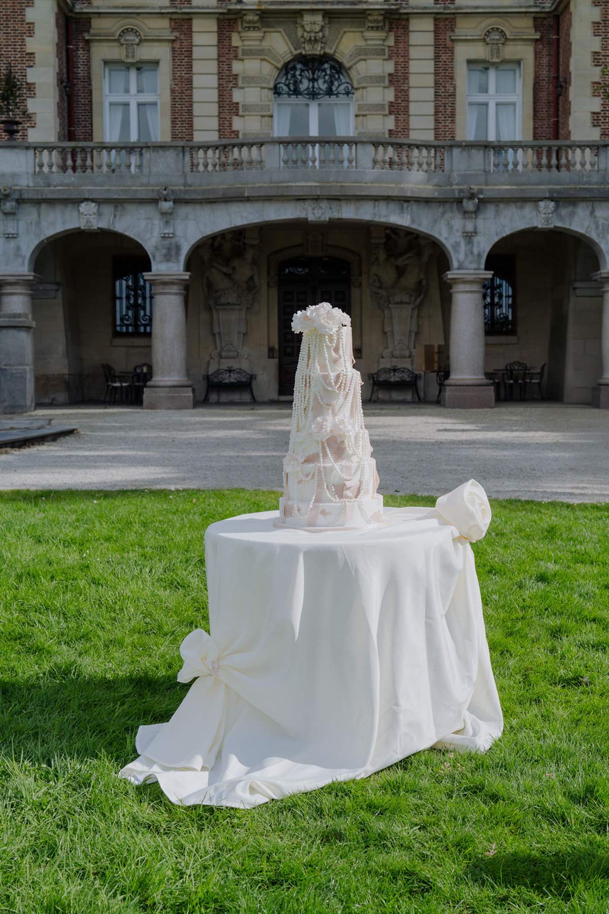 Multi-tiered blush cake with pearl draping and sugar butterflies before red brick chateau colonnade