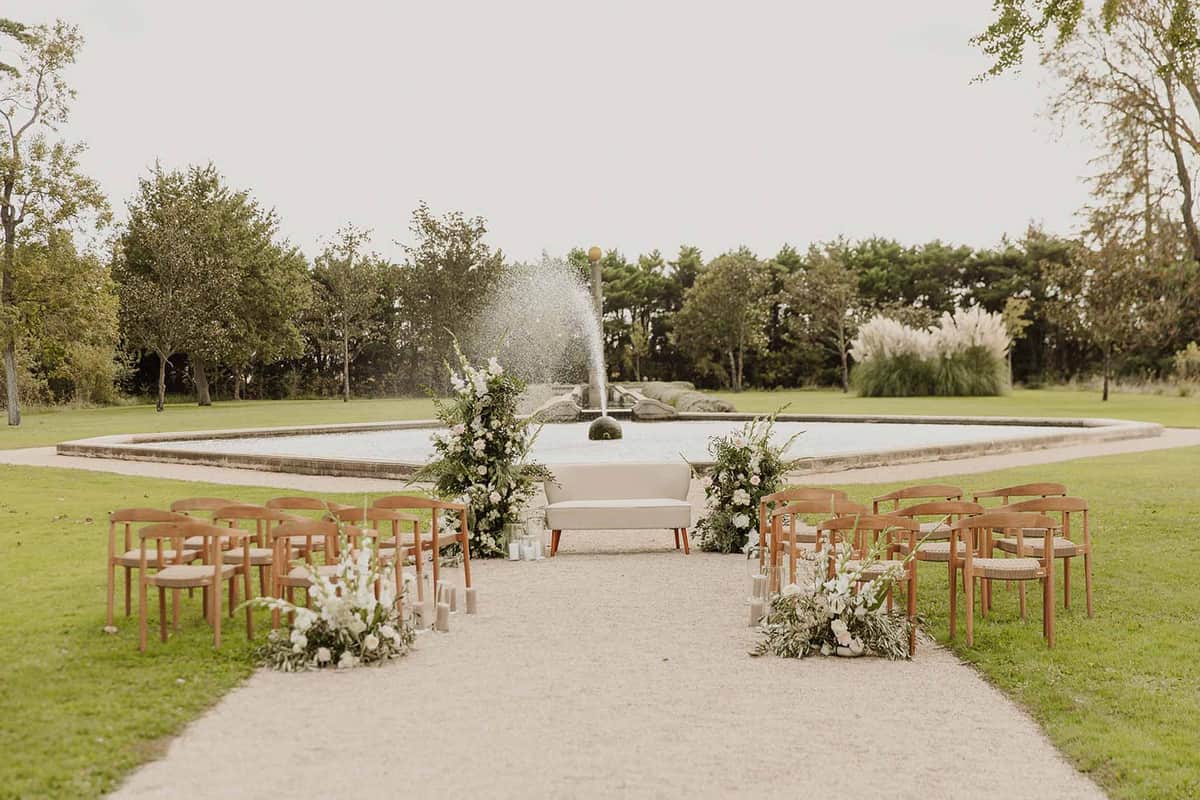 Ceremony aisle with wood chairs white and blush florals leading to cream sofa altar before stone fountain