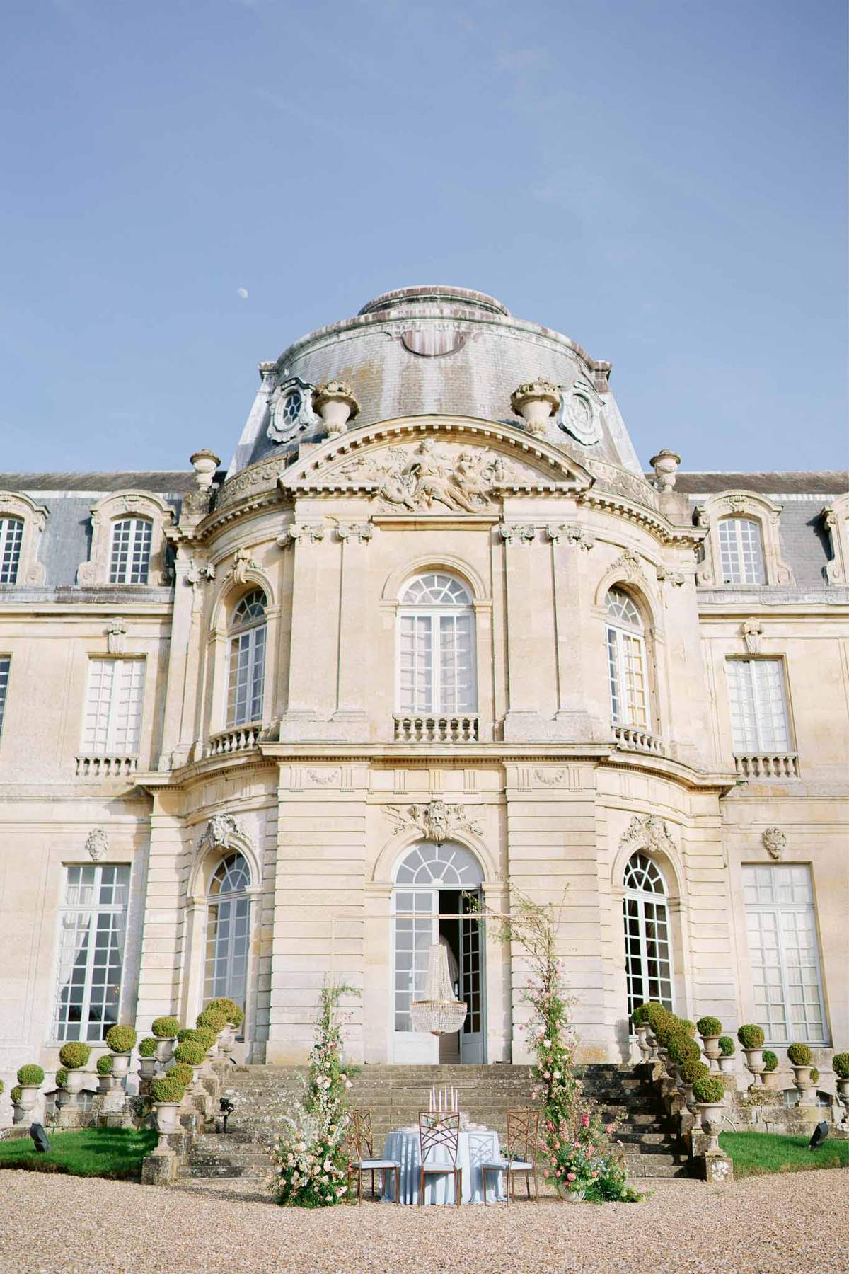 Baroque chateau facade with sweetheart table on forecourt, blush floral installations flanking entrance