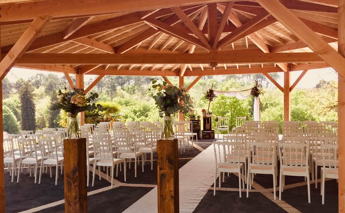 Wooden pavilion ceremony space with white chairs, floral aisle arrangements, and sheer fabric backdrop before guests arrive