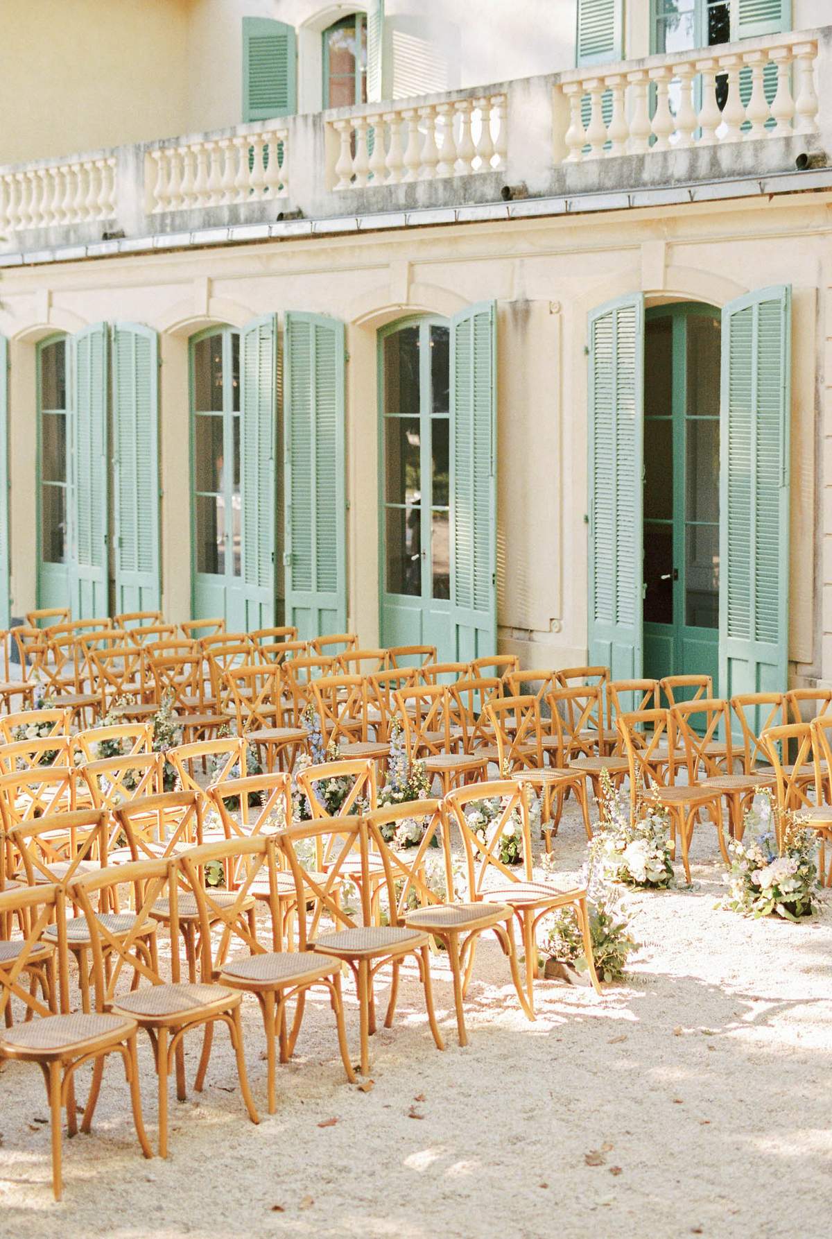 Ceremony setup with cross-back chairs and blush blue aisle florals before sage-shuttered chateau