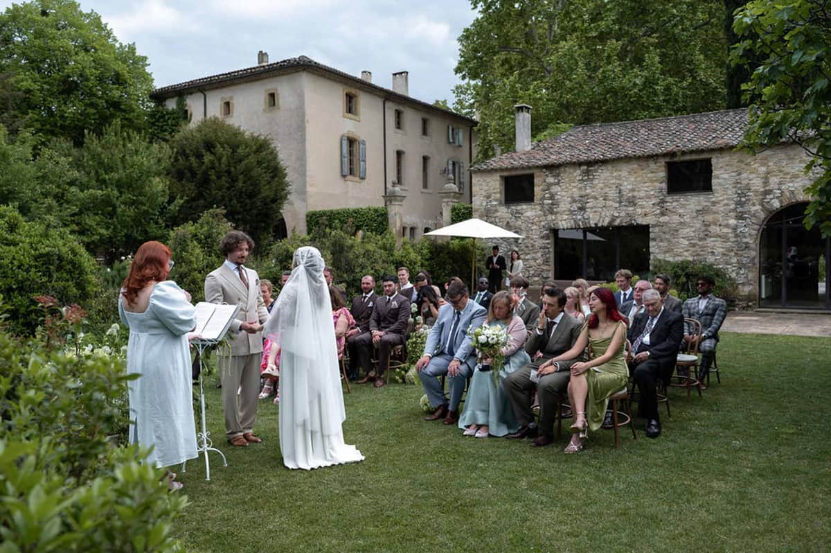 Outdoor lawn ceremony before country estate with blue shutters and guests on bistro chairs
