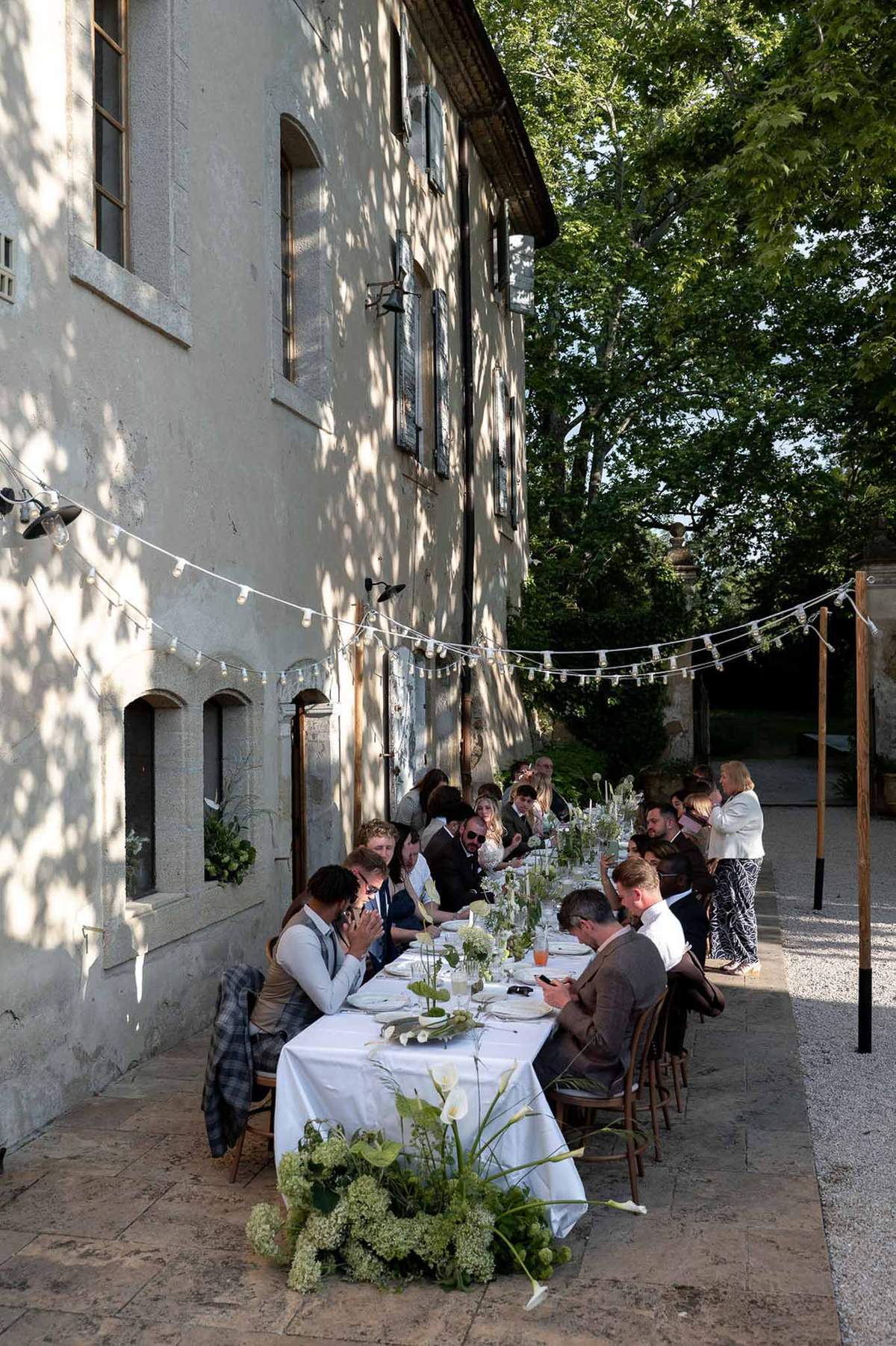 Twenty-five guests at banquet table with calla lily runner bistro lights and bentwood chairs along manor facade