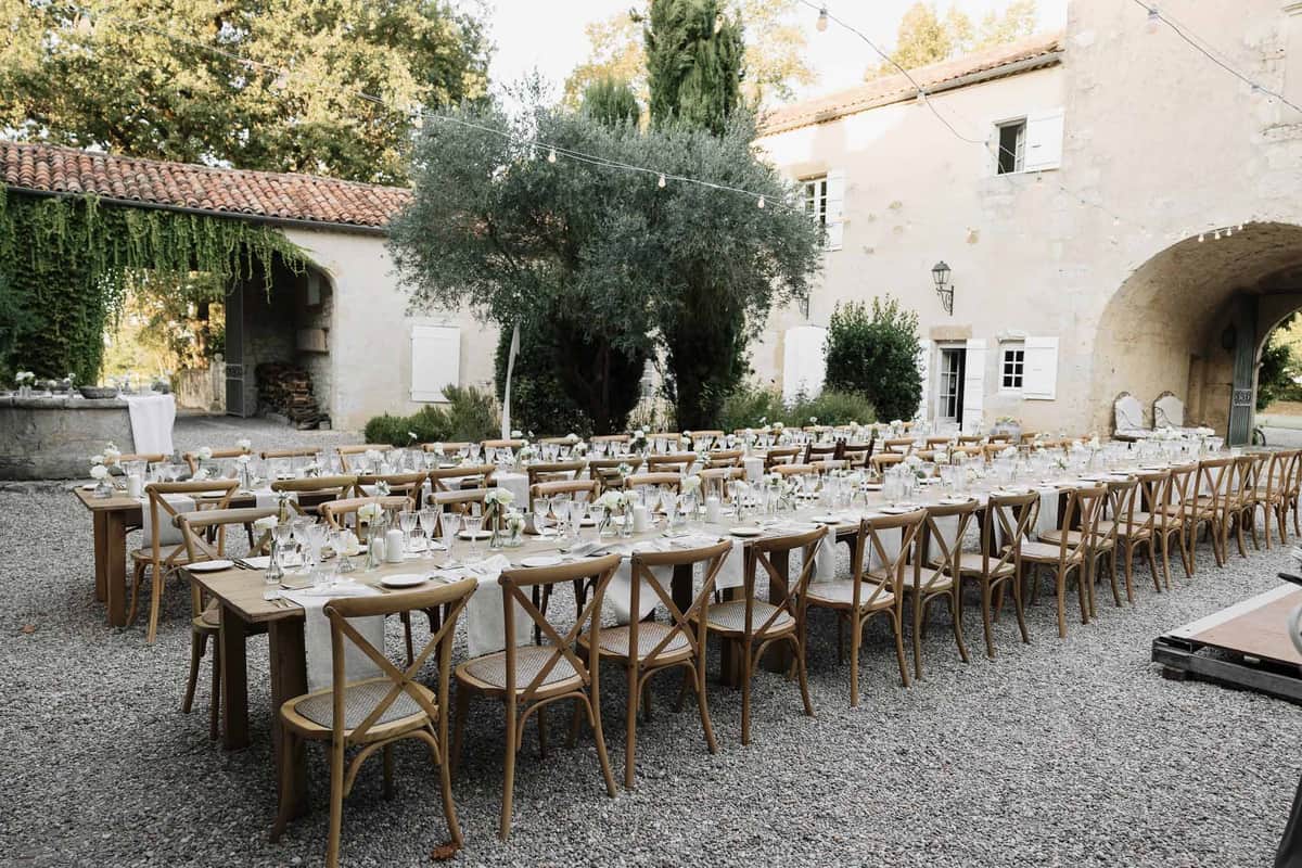 Outdoor reception setup in a Provencal courtyard with long farm tables, cross-back chairs, white florals, and overhead fai...