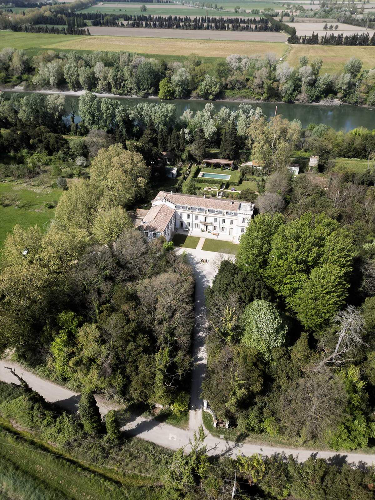 Aerial view of French chateau with terracotta roof, swimming pool, formal gardens, and river alongside