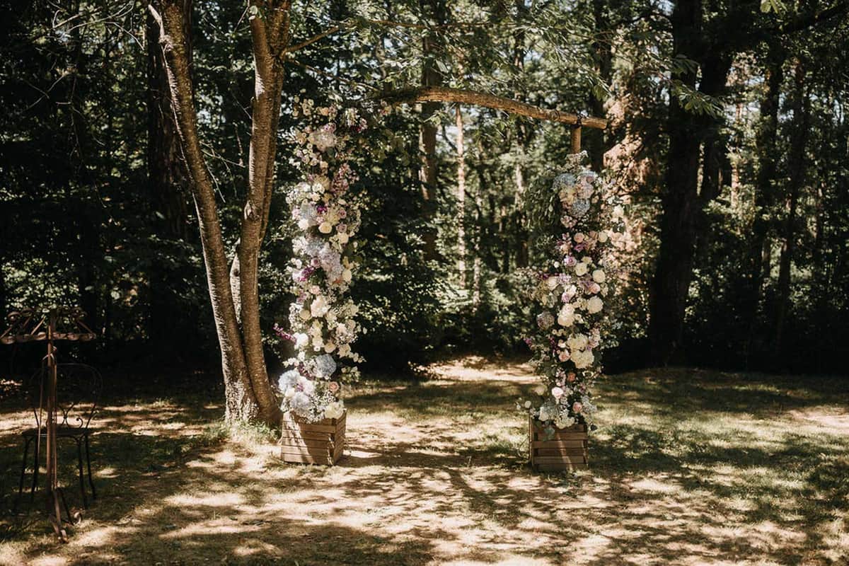 Rustic wooden ceremony arch decorated with ivory hydrangeas, blush roses, and mauve blooms in a sunlit forest clearing