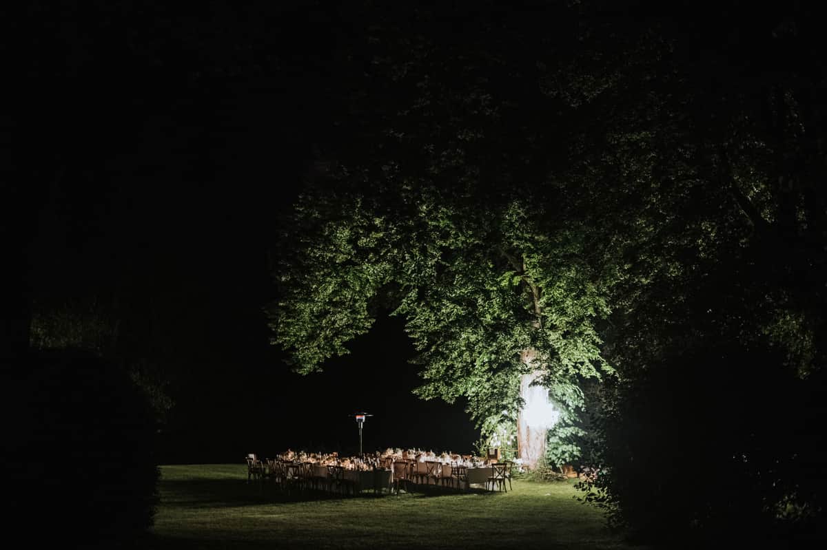 A wide-angle nighttime shot of an outdoor wedding reception dinner set up on a lawn, with multiple long rectangular tables arranged in a U or parallel formation, dressed in white linens and set with glassware, candles, and floral centerpieces in warm amber tones. The tables are surrounded by cross-back wooden chairs and lit by warm candlelight and what appears to be an overhead chandelier or hanging light installation suspended from a large mature tree, which is dramatically uplighted in green. The surrounding grounds are dark, with the illuminated table setting and tree forming a focal point against the black night sky. No guests are visible, suggesting the shot was taken before or between service. The composition is a distant wide shot taken from ground level across the lawn, framing the scene through the dark silhouette of hedgerows.