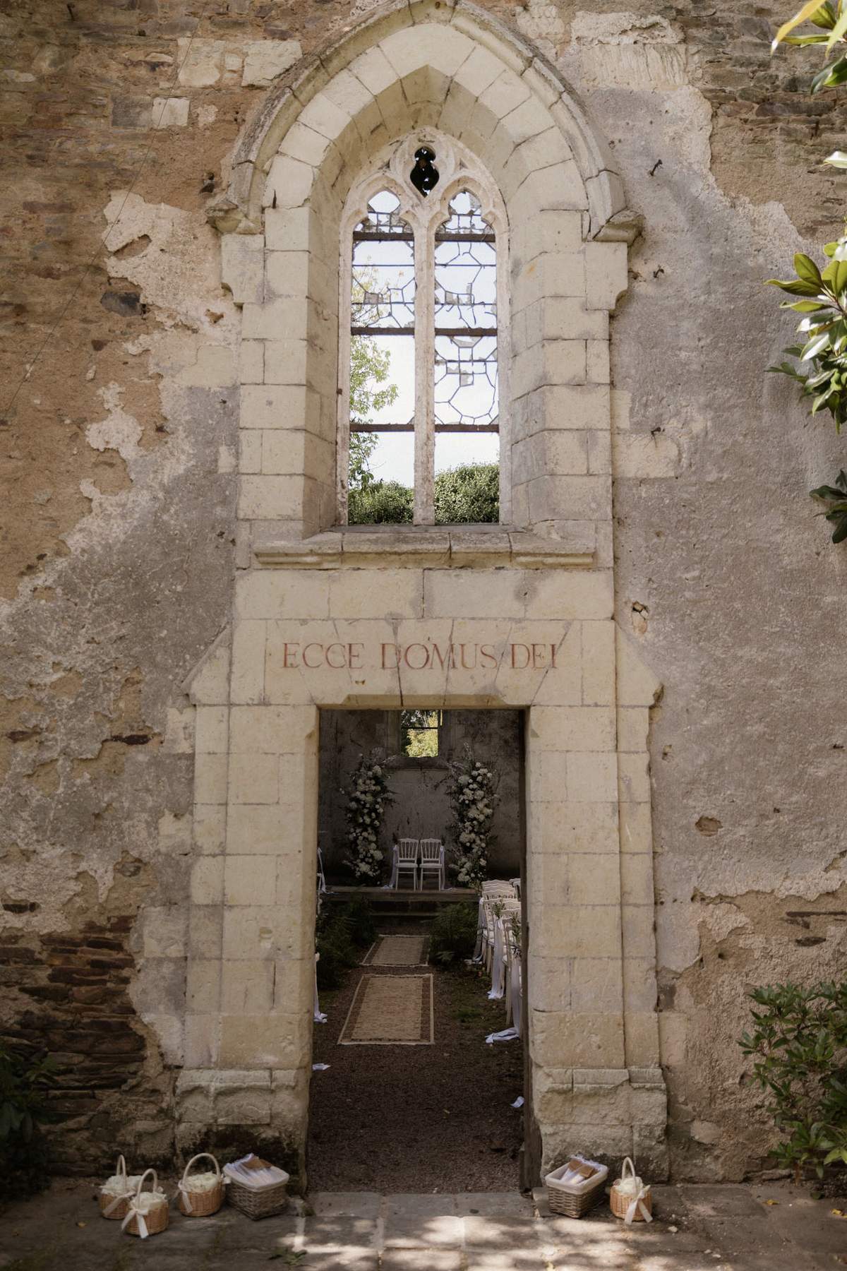 The exterior entrance of a partially ruined stone chapel is shown set up for a wedding ceremony, viewed from outside through the doorway. The Gothic-arched stone portal bears the Latin inscription 'ECCE DOMUS DEI' above the entrance, and above that sits a Gothic lancet window with remnants of leaded glass. Through the doorway, a natural jute aisle runner leads toward an altar area flanked by two tall white floral columns — likely composed of white blooms and greenery — with a small arrangement of white chairs set for the ceremony and guest seating visible on either side of the aisle. Wicker baskets containing white flower petals or confetti are placed at the base of the entrance on either side. The overall decor palette is white and natural/neutral, with a classic romantic styling suited to the historic architectural setting. Wide shot capturing the full facade and ceremony setup within. Potential venue feature image.
