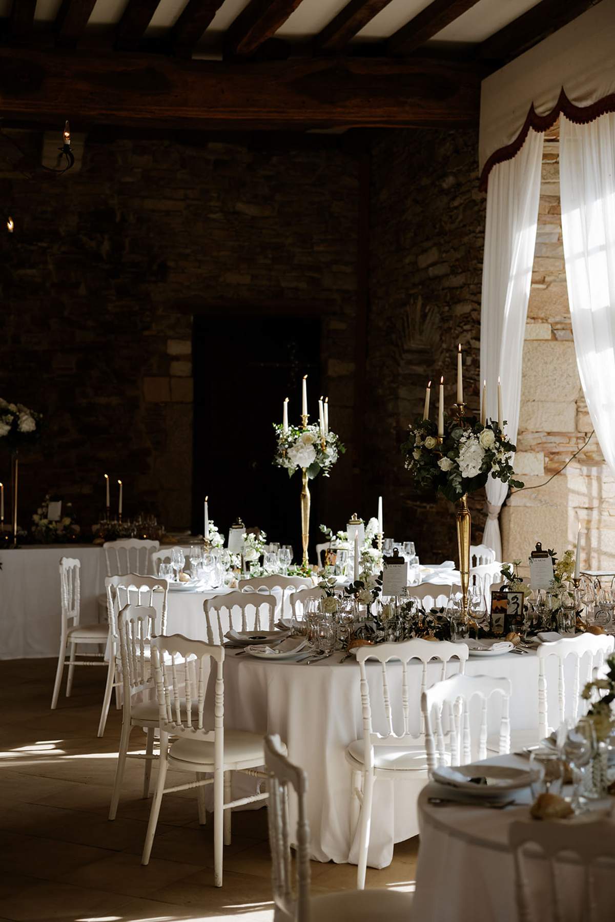 Indoor wedding reception setup photographed in a wide shot, showing round tables dressed in white floor-length linen cloths inside a stone-walled room with dark exposed wooden ceiling beams. The tables are surrounded by white Napoleon chairs and set with glassware, grey linen napkins, and place settings. Tall gold candelabras hold lit ivory taper candles and are topped with white floral arrangements featuring white roses, white hydrangeas, and greenery including eucalyptus. A trailing greenery and white floral runner decorates the table surface. Sheer white curtains with a burgundy scalloped trim hang at the window, allowing natural light to flood the space. Additional candlelit tables are visible in the background, maintaining the same white and gold decor palette. The overall styling theme is classic and formal with a green and white floral palette against gold metallic accents.