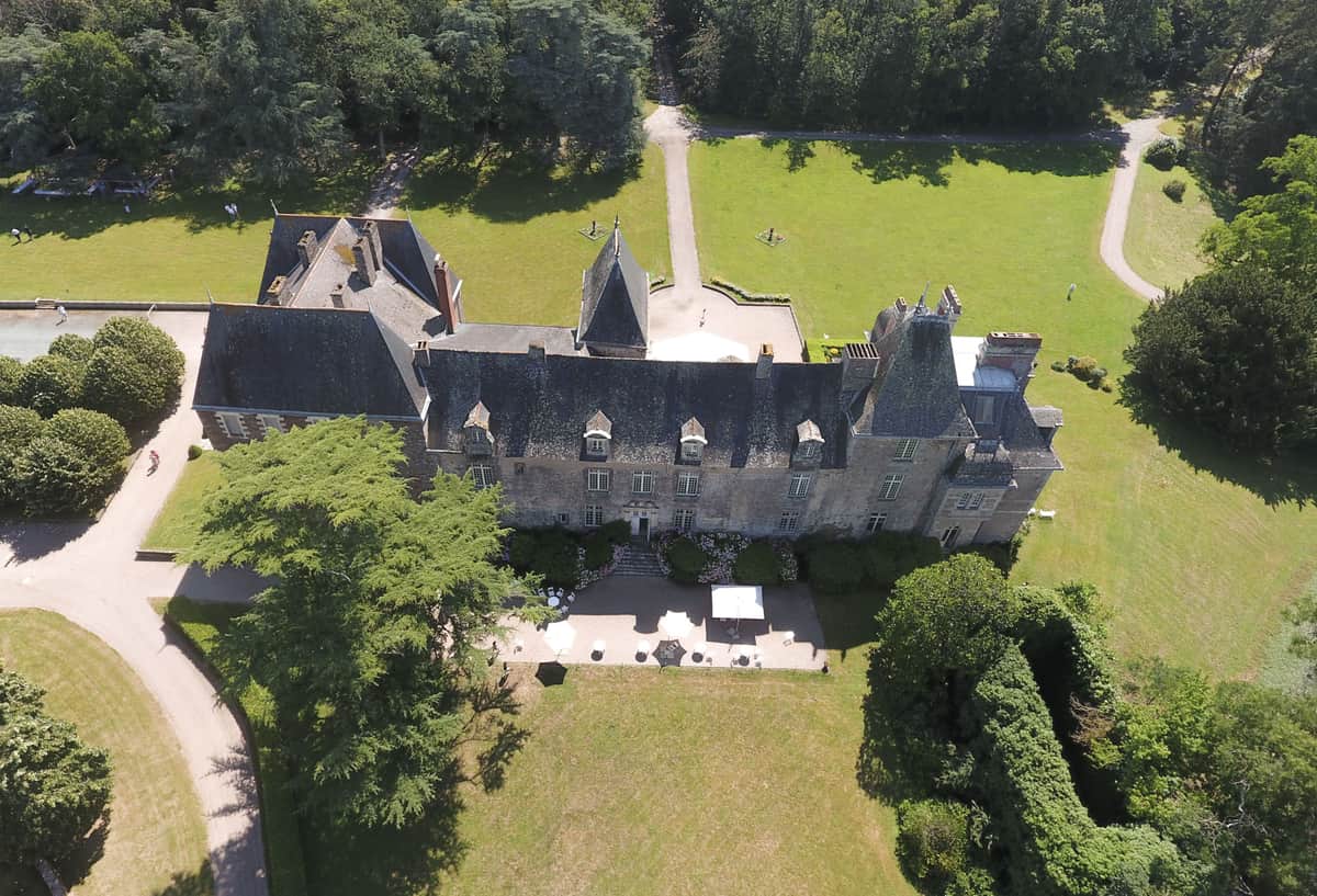 An aerial wide shot of a French château wedding venue, showing the full stone manor house with dark slate mansard roofing, pointed turrets, and dormer windows characteristic of French Renaissance architecture. In front of the château's main entrance, white market umbrellas and what appear to be white-draped tables are set up on a terrace courtyard, indicating an outdoor cocktail hour or reception in progress, with a small number of guests visible around the setup. The property is surrounded by extensive formal grounds including wide lawns, gravel driveways, clipped ornamental trees, and dense woodland at the perimeter. Potential venue feature image.