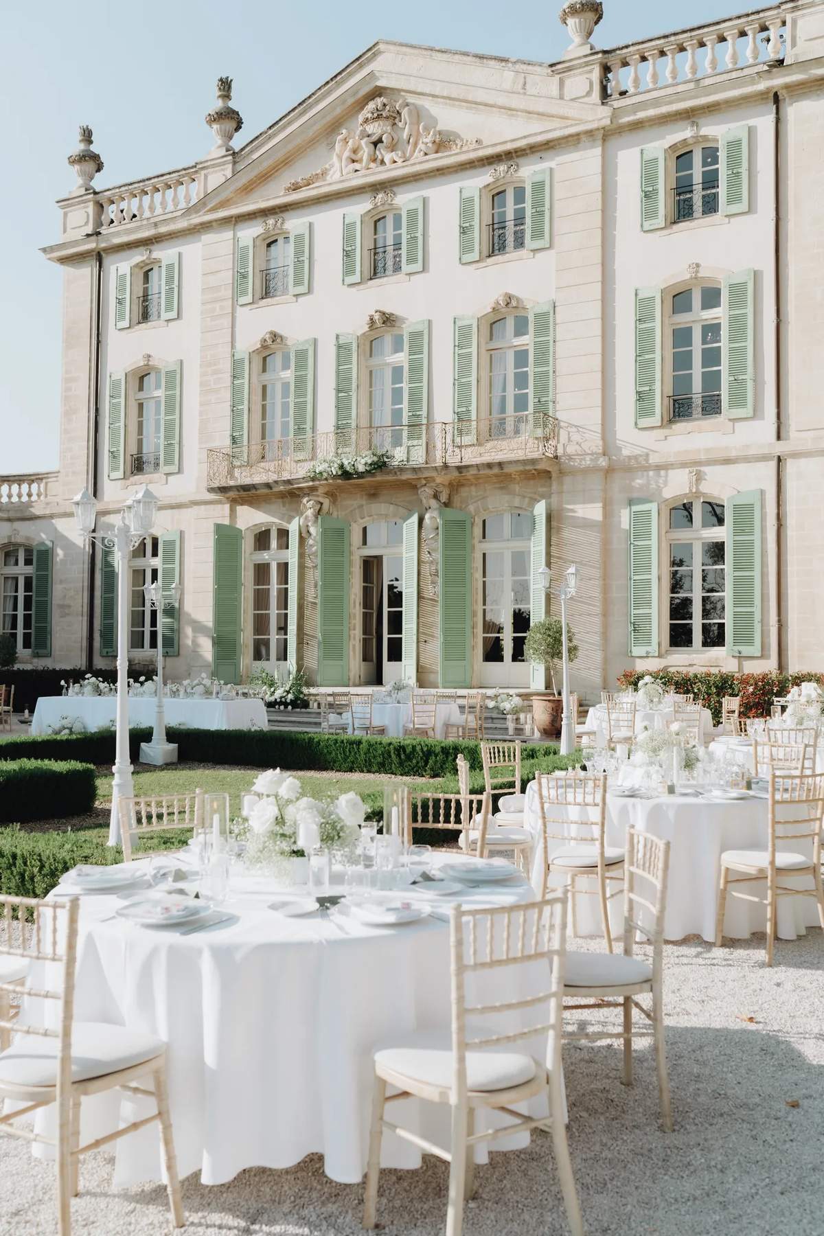 Round wedding tables on the gravel terrace of a château with pale green shutters, by Be My Love Weddings