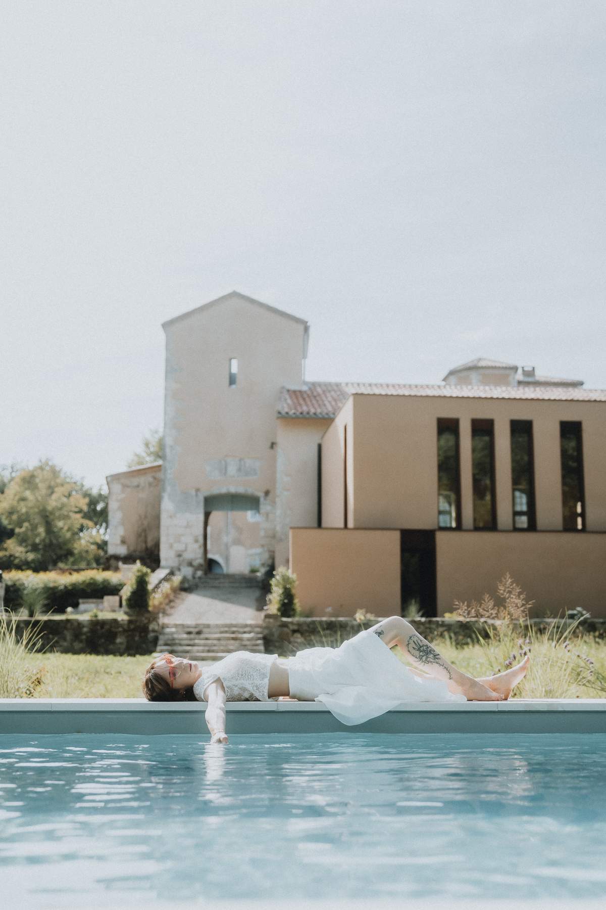 A bride in a white dress reclining at the edge of the swimming pool, with the château's modern extension building behind.