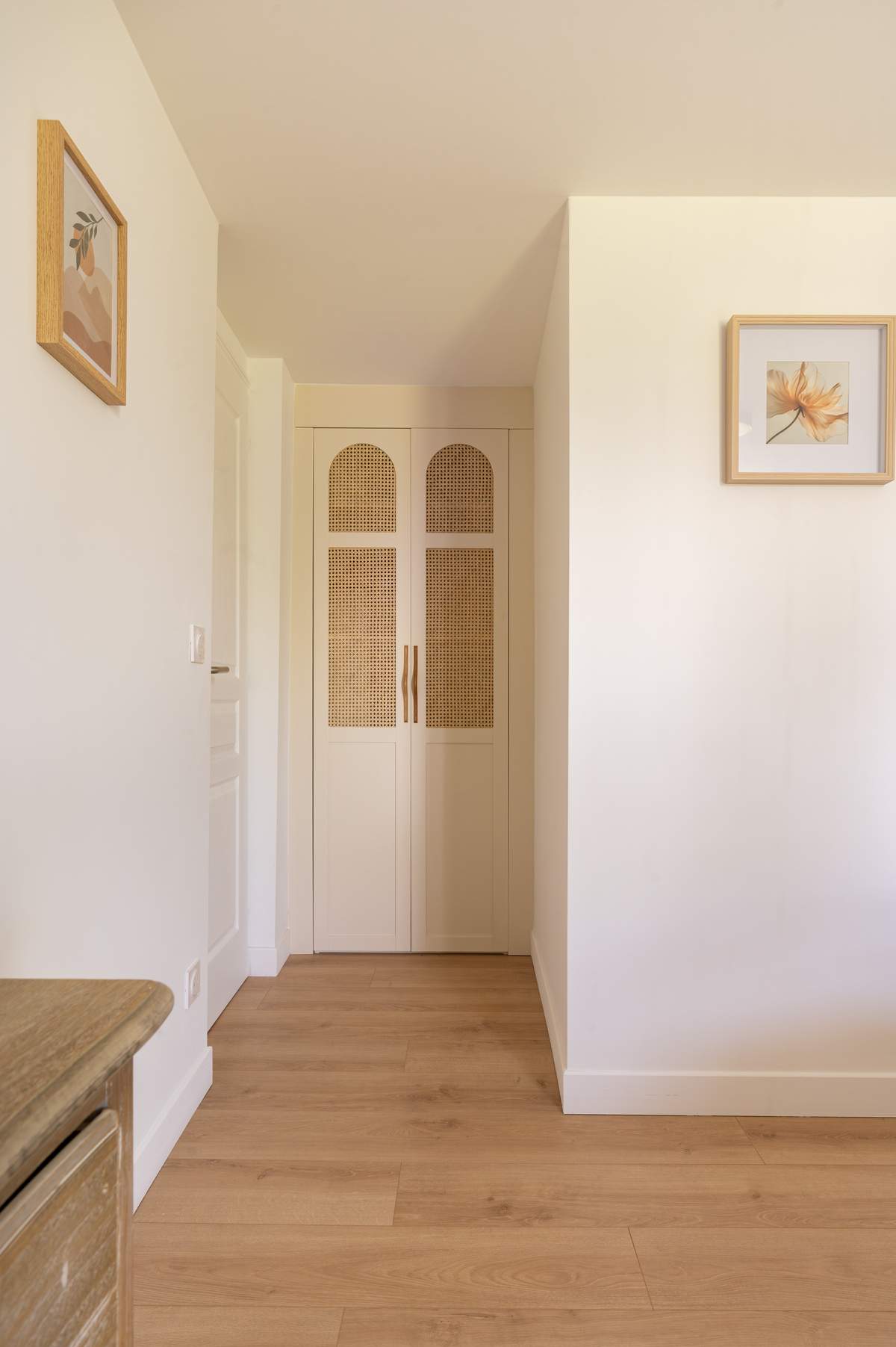 A hallway with arched cane wardrobe doors, oak flooring, a framed botanical print and a figure study