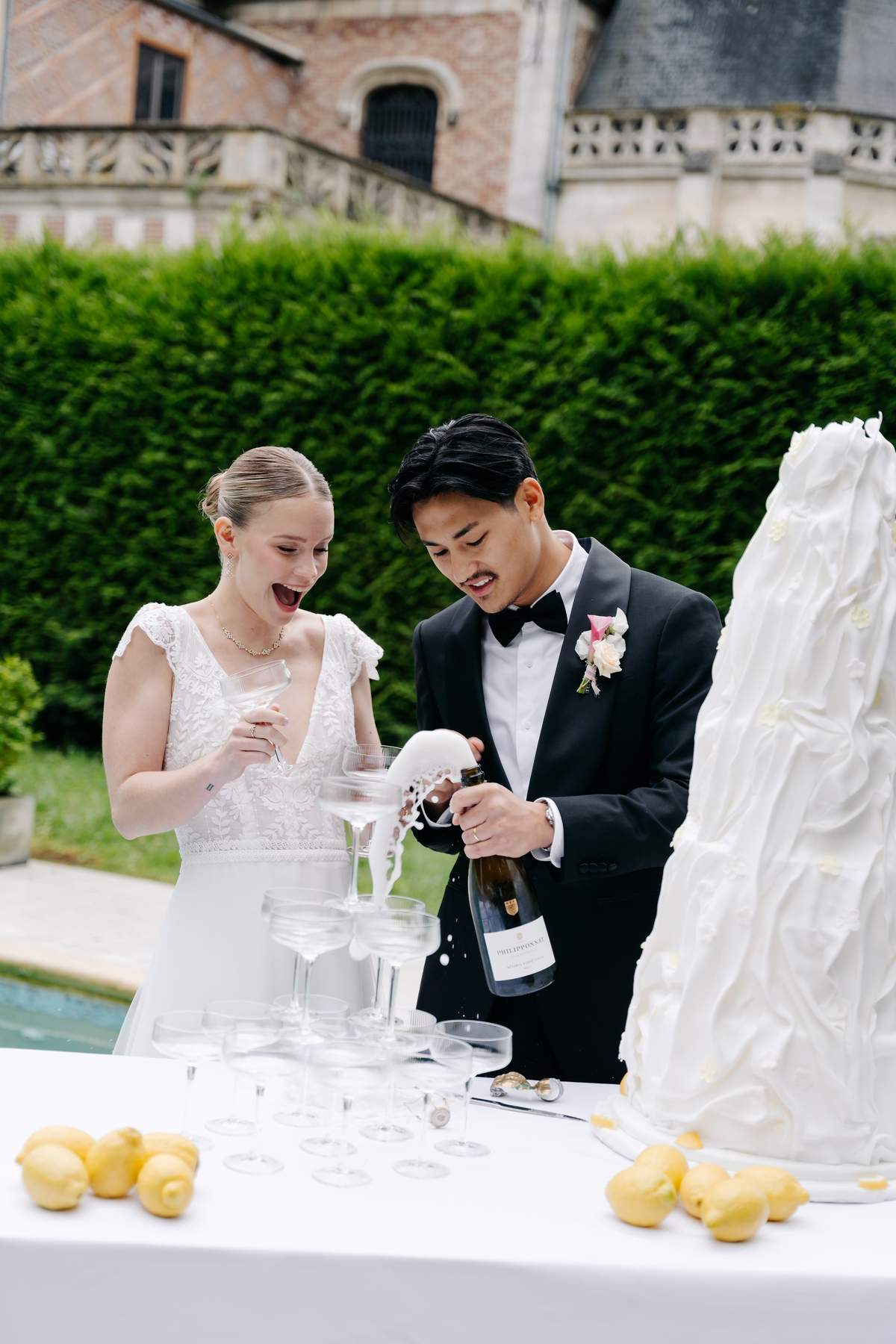 A bride laughing, coupe in hand, as the groom pours champagne over a coupe tower by the pool, beside a tall white cake