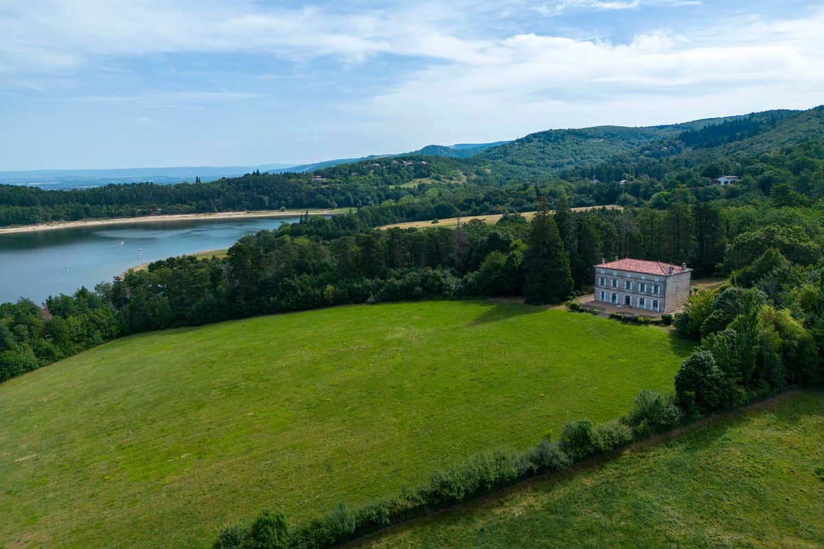 Aerial view of the château beside its meadow above the lake, with wooded hills beyond