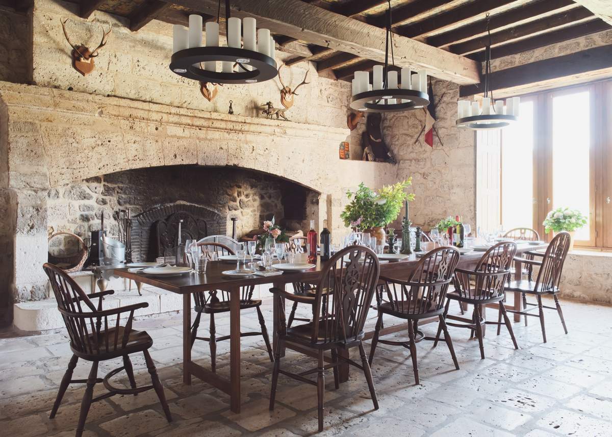 Long dining table set with glasses under candle chandeliers, beside a vast stone fireplace and mounted antlers