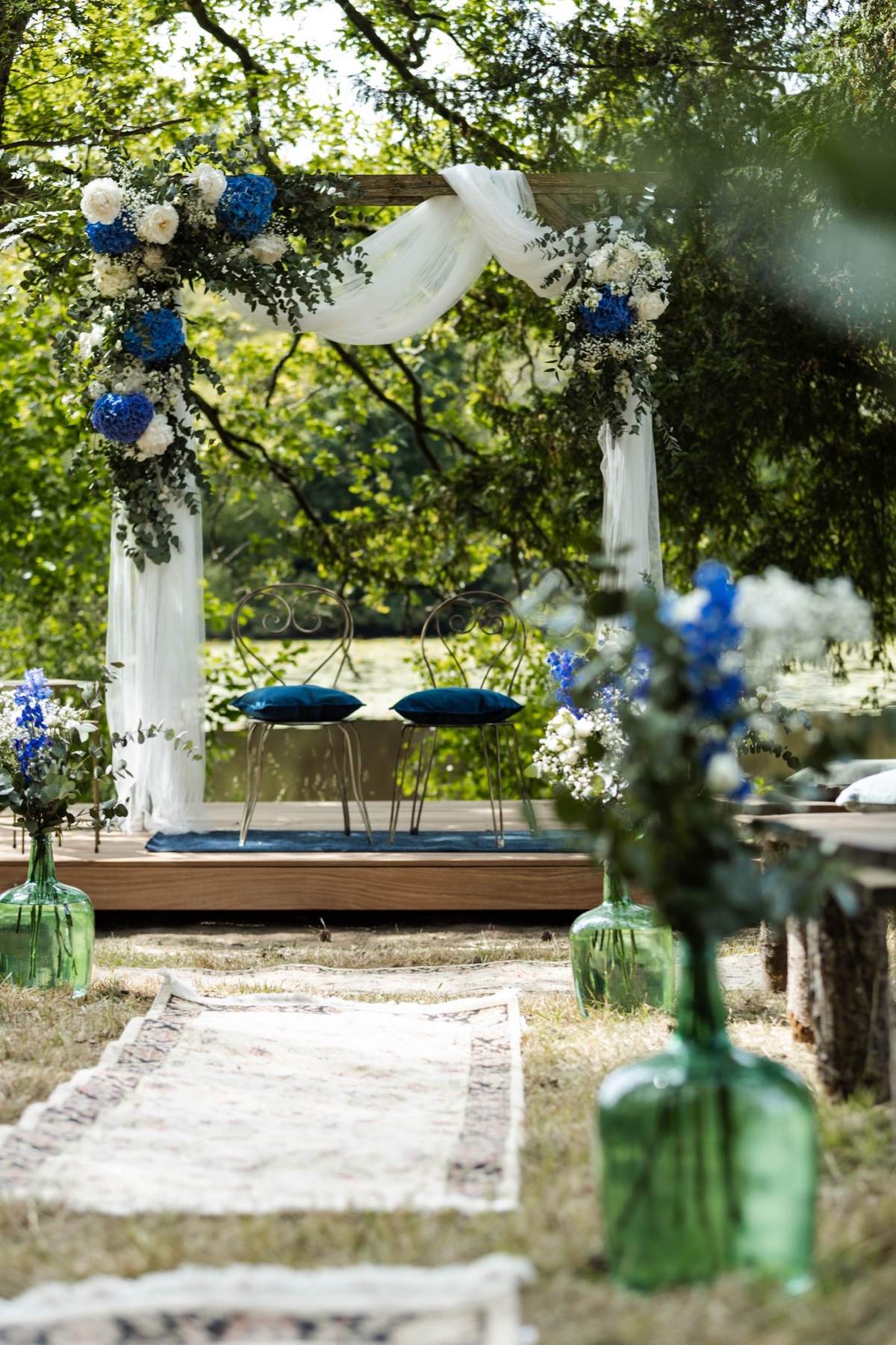 Outdoor ceremony arch at Chateau du Pordor dressed with blue and white flowers, with an aisle runner and green demijohns