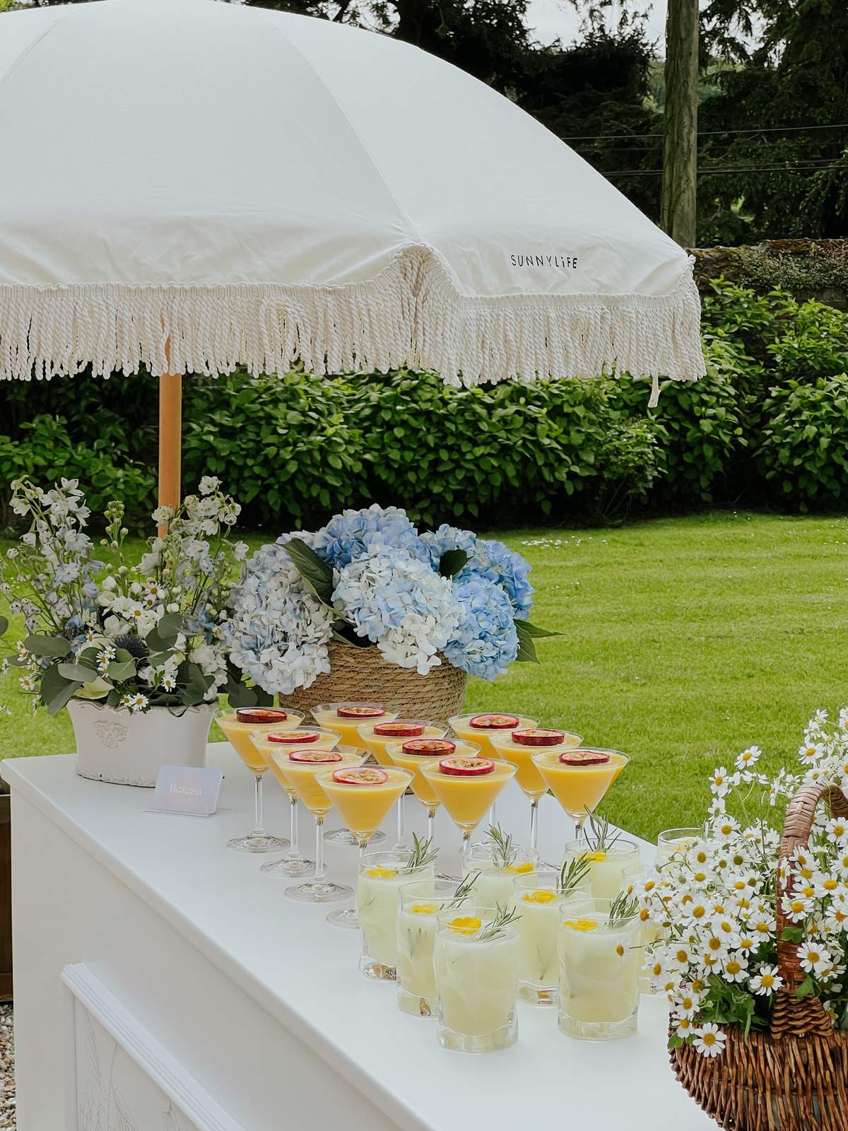 A drinks table set with cocktails and hydrangeas under a fringed parasol on the lawn