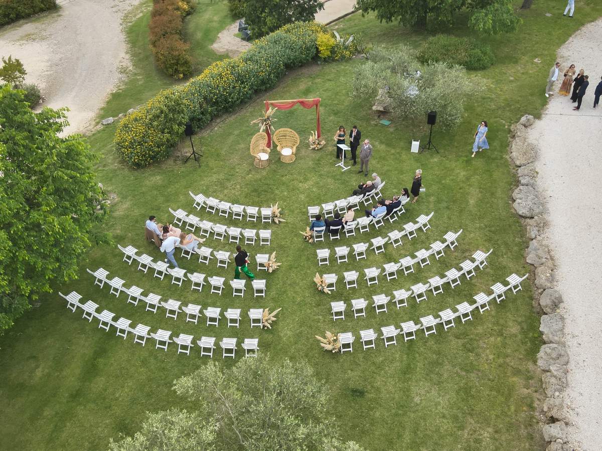 Aerial view of a lawn set with curved rows of white folding chairs facing a draped arch, with two wicker chairs and a lectern beside a flowering hedge.